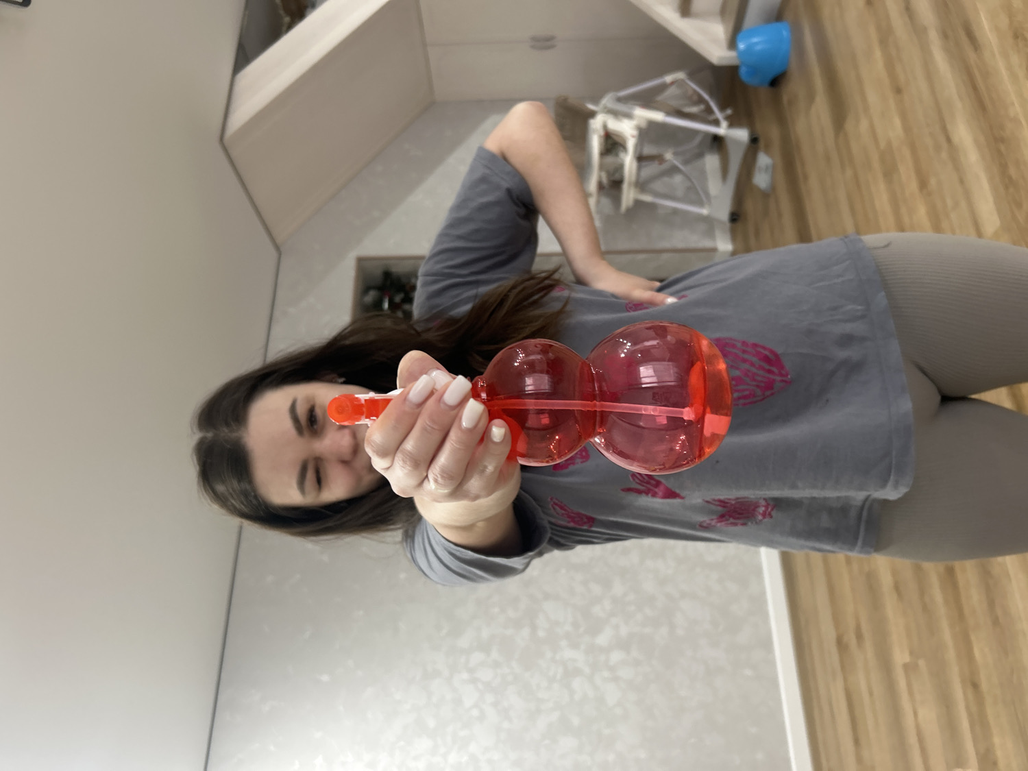 The image captures a young woman indoors, playfully pointing a red, translucent, double-bulb-shaped spray bottle directly towards the viewer. She has dark hair, a light complexion, and is winking or squinting with a mischievous expression. Her fingernails are painted with white polish. She is wearing a grey t-shirt adorned with a pink graphic design and light grey or beige leggings.
The setting appears to be a domestic room, possibly a living area or bedroom, with light-colored walls and a light wooden floor. In the background, to her right, a light-toned piece of furniture, perhaps a shelf or desk, is visible. Further back, there's what looks like a white baby bouncer or playpen, and a bright blue bowl or container rests on the wooden floor. The room is well-lit, suggesting either daytime or bright interior lighting. The overall mood of the image is lighthearted and playful. The scene is set in Chaikovsky, Russia. There is no visible text.
