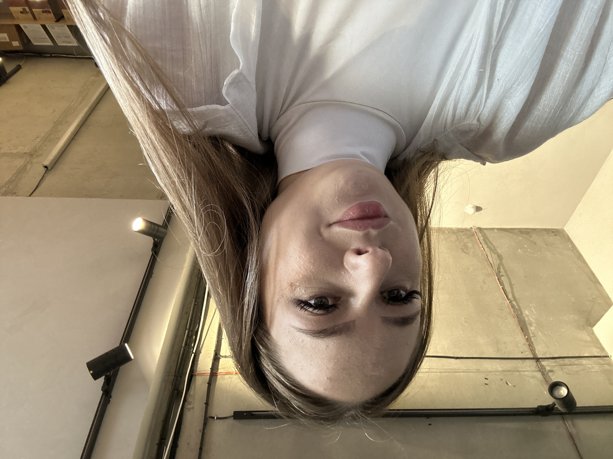 A young woman with long brown hair is pictured from the shoulders up, looking directly at the camera. She is wearing a white turtleneck shirt and a sheer white open-front shirt. The background appears to be an indoor space with a concrete ceiling and exposed lighting fixtures. The lighting is overhead and casts shadows. The woman is looking slightly downwards.