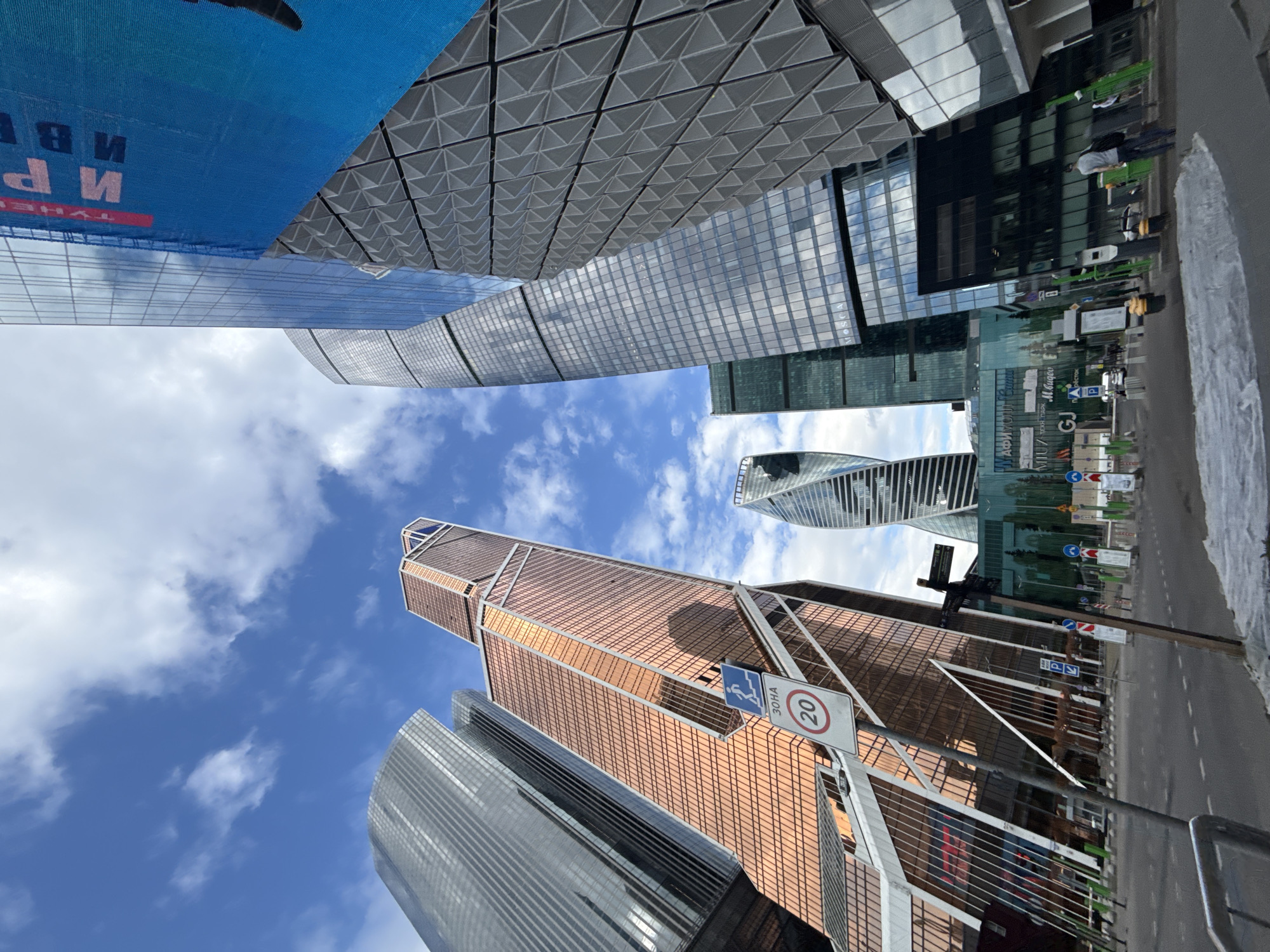 The image shows a view looking up at several modern skyscrapers in the Moscow International Business Center (Moscow City) district. The sky is blue with scattered white clouds. A speed limit sign with the number "20" is visible. Street signage and road markings are present on a roadway to the right. A blue banner is seen in the top left corner. There is no indication of Khimki, Russia.