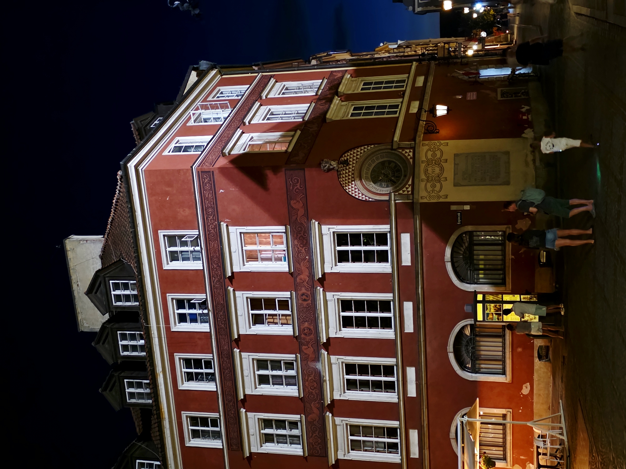 This is a nighttime exterior shot of the tenement at 37 Rynek Starego Miasta in Warsaw, Poland, part of the Old Town Market Square. The building is painted a deep red with white trim and adorned with decorative friezes featuring patterned designs and winged figures. Multiple floors of windows with white frames are visible, some open, others closed, with a few illuminated from within. A prominent circular clock face is mounted on the corner of the building, above an arched recessed area. Several people are present on the cobblestone street. In the foreground, a group of individuals stand near a lit information kiosk. Further down the street, other figures are walking, with streetlights and building lights illuminating the scene against a dark blue night sky.