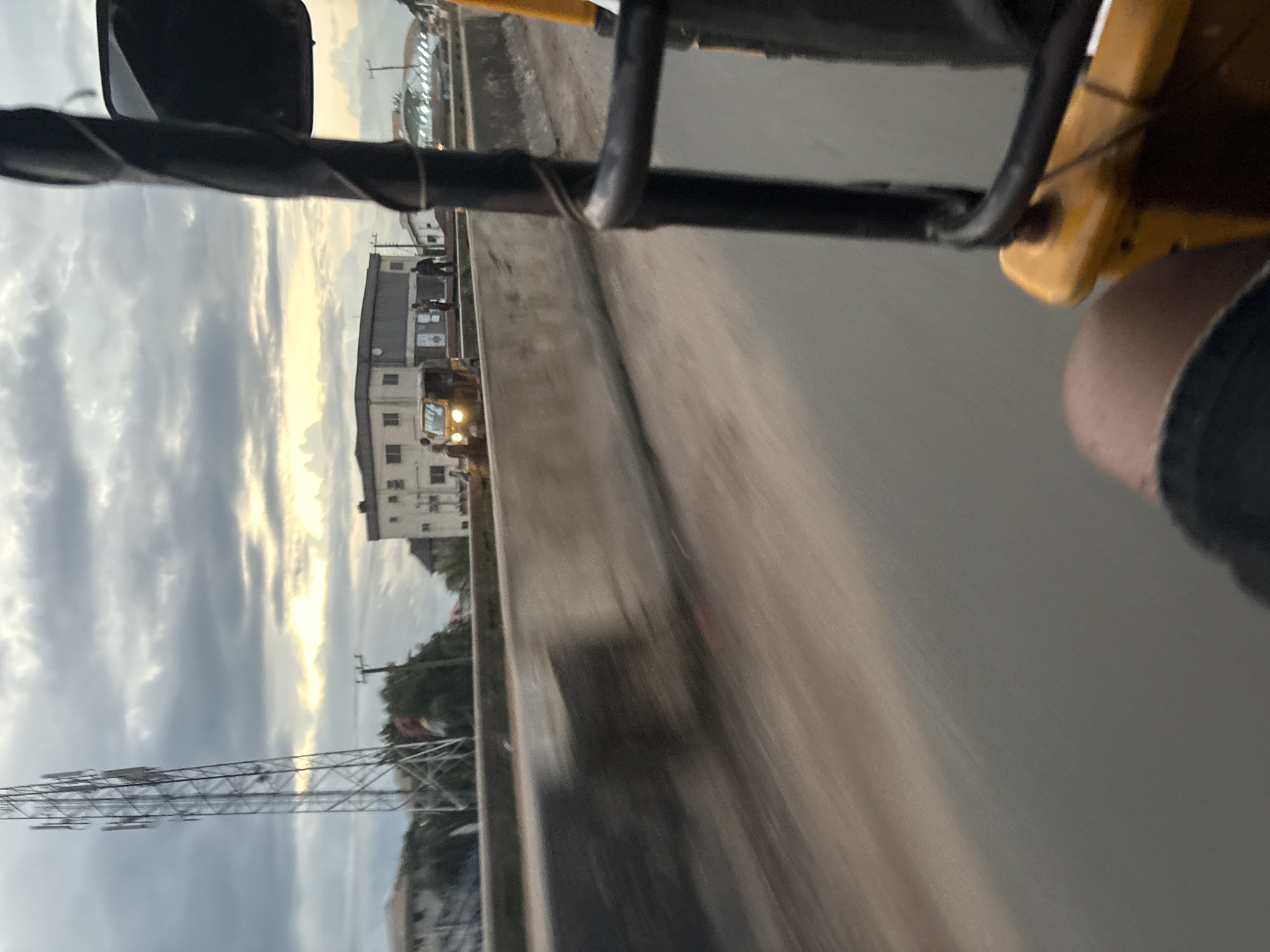 The image depicts a view from a moving vehicle, likely a tricycle, on a road in Onitsha, Nigeria. A concrete barrier runs along the side of the road, with blurred motion suggesting speed. In the background, a telecommunications tower stands tall against a cloudy sky with hints of sunlight breaking through. Buildings, including a multi-story structure and smaller houses, are visible beyond the barrier. Several people are standing near a yellow tricycle on the road. The scene suggests a typical urban streetscape during the day.