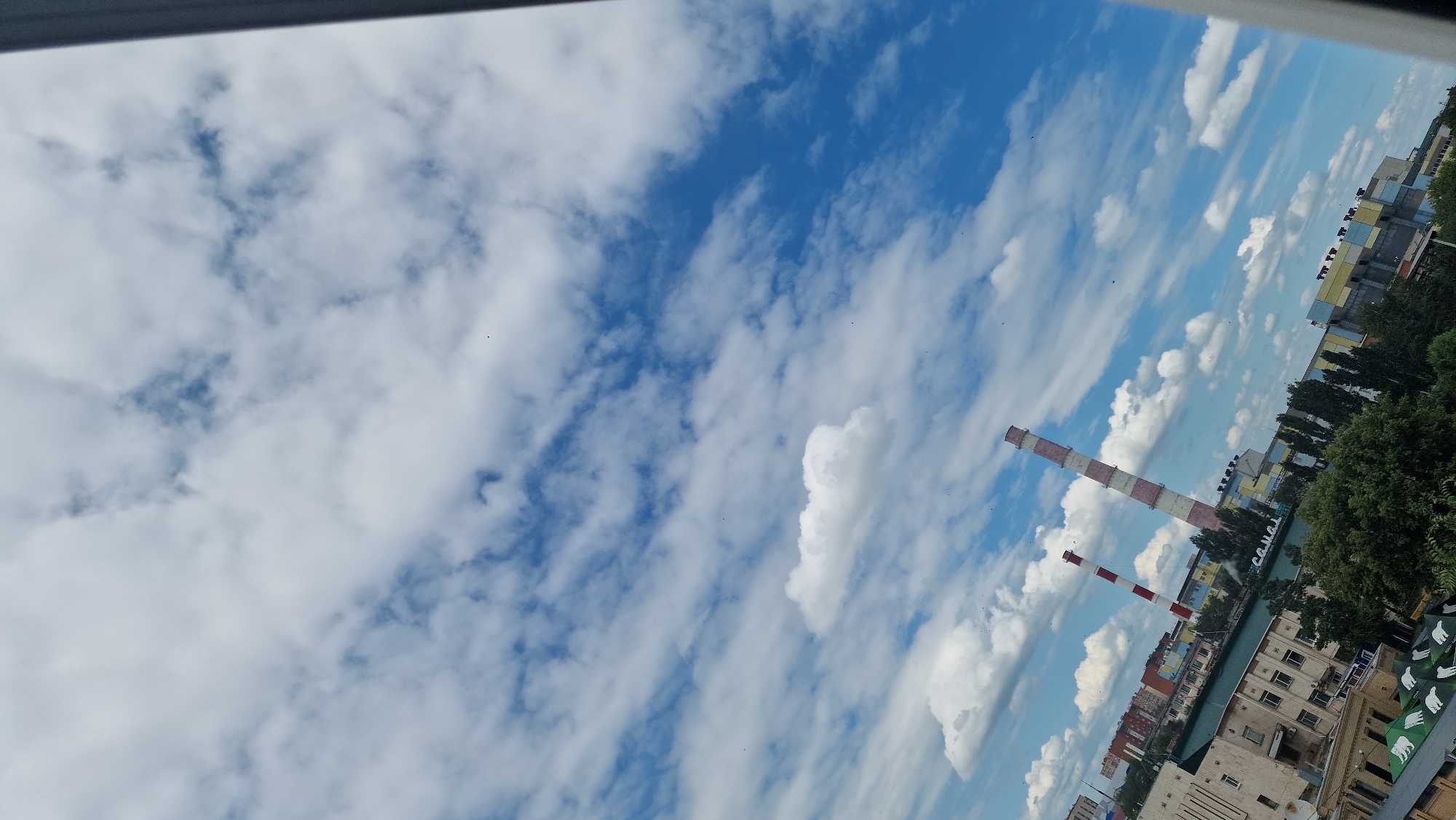 A medium-angle shot captures a cityscape under a partly cloudy sky. Two tall industrial smokestacks, one with red and white stripes and the other with a more faded red and white pattern, dominate the middle ground, pointing diagonally upwards towards the sky. Behind the smokestacks, a collection of buildings can be seen, some appearing residential and others more industrial with colorful facades. In the foreground, the tops of green trees and some structures with what appear to be umbrellas are visible, suggesting a viewpoint from a higher elevation. The sky is a vibrant blue, filled with scattered white clouds that vary in size and density. The image is oriented vertically, with the top of the frame primarily filled with sky. The location appears to be the industrial zone near the Tobol River in Kostanay, Kazakhstan, specifically the area surrounding the Kostanay Thermal Power Plant (TETS-2).