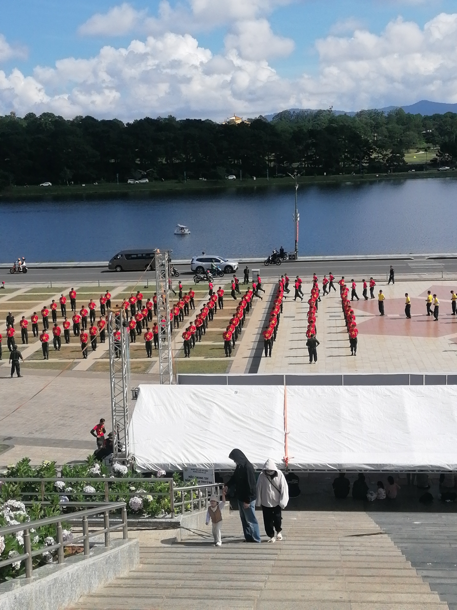 The image depicts a wide-angle view of an outdoor event in Da Lat, Vietnam, likely at Xuan Huong Lake, given the presence of a large body of water and a treelined embankment. In the foreground, a set of concrete stairs leads down to a paved area where a large gathering is taking place. A group of people, predominantly young males dressed in red shirts with yellow stars and dark pants, are arranged in formation on the pavement. Some are standing, while others appear to be moving or in a synchronized pose. A few individuals in yellow shirts and dark pants are also present within the formation.
Further back, a large white tent structure is set up, likely for seating or a stage. Near the tent, several individuals are seated on the ground. Two people, one wearing a dark hoodie and the other a white hoodie, are walking down the stairs in the foreground. A young child in a light-colored outfit is also seen walking down the stairs.
Beyond the paved area, a road runs alongside the lake. Several vehicles, including a silver van, a white SUV, and motorcycles, are parked or moving on the road. The lake itself is calm and reflects the blue sky with scattered white clouds. A small white boat is visible on the water. The background features a dense line of dark green trees, beyond which rolling hills and mountains are faintly visible. The overall scene suggests a public event or performance.