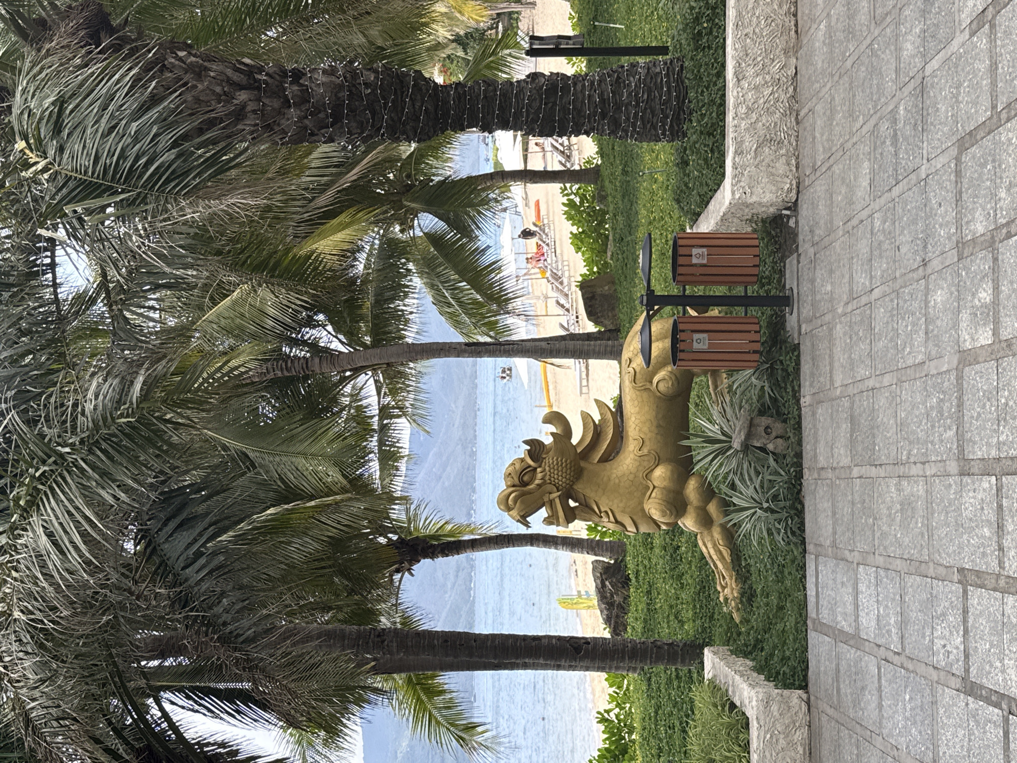 The image shows a beachfront scene in Nha Trang Ward, Vietnam. A golden dragon statue stands prominently on a grassy strip between a paved walkway and the beach. Palm trees are visible throughout the frame. In the background, the beach is visible, featuring lounge chairs, umbrellas, and people. The ocean extends to the horizon. Two trash receptacles are also visible.