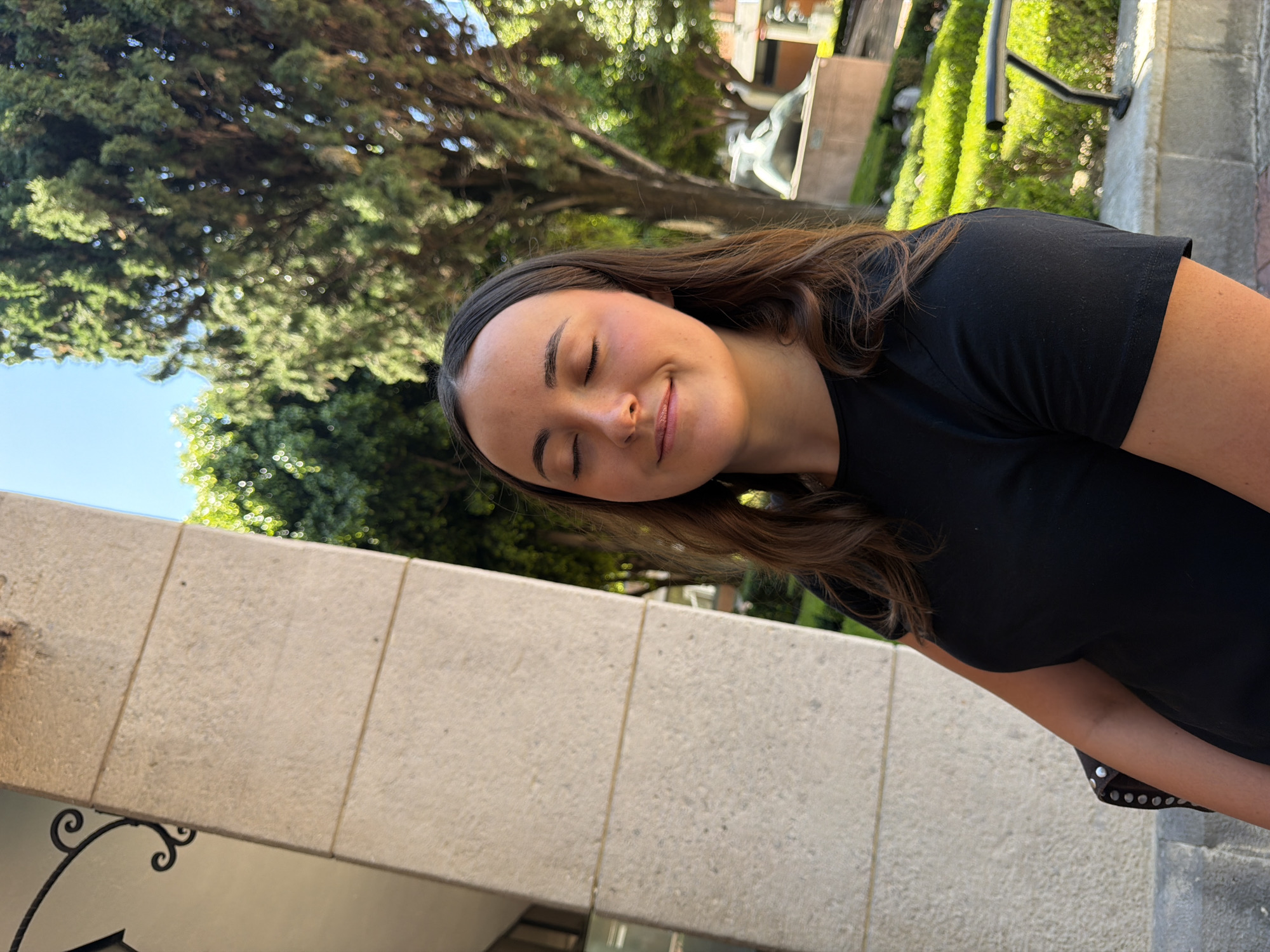 A slightly tilted, outdoor portrait captures a young woman with long, dark brown hair, smiling gently with her eyes closed, conveying a sense of peace and contentment. She is wearing a simple black t-shirt. The bright natural light suggests it's daytime, under clear skies visible in the upper left.
The setting appears to be a well-landscaped outdoor area, possibly a park, garden, or courtyard in San Andrés Cholula, Mexico. Lush green trees with blurred foliage dominate the background, particularly in the upper left, creating a natural canopy. Architectural elements are visible: a light-colored, possibly stone or concrete, wall or ledge with vertical seams runs along the bottom and left side of the frame. Behind the woman, in the mid-background, indistinct structures and a blurry, light-colored statue or sculpture can be seen amidst more greenery and what appears to be a dark metal railing. The shallow depth of field keeps the woman in sharp focus while blurring the background elements. There is no visible text in the image.