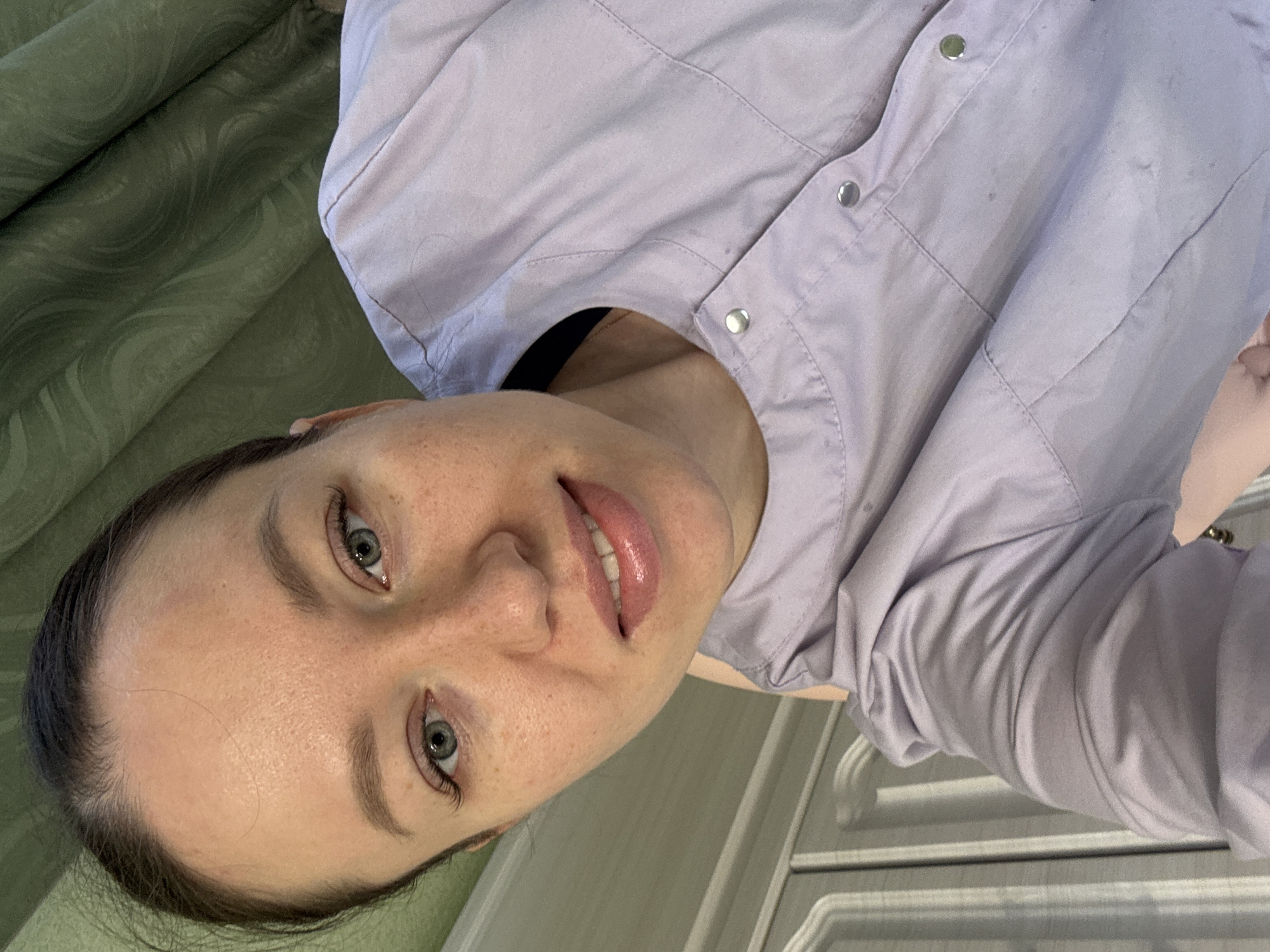 The image displays a close-up, self-portrait of an adult female located in the city of Ферма, Russia. The woman has light skin with scattered freckles visible on her nose and cheeks. Her dark brown hair is pulled back, revealing her forehead. Her eyes are light green, and her lips, coated with a glossy, light pink substance, are slightly parted in a smile, revealing her upper teeth. She wears a light purple, collared top, possibly a uniform or smock, fastened with silver snap buttons. A dark-colored strap is visible beneath the collar on her left side. The background behind her upper left consists of a dark green, patterned fabric, consistent with a curtain. To her lower left, a light-colored furniture piece with vertical paneling and molding is partially visible. The woman is looking directly into the camera.