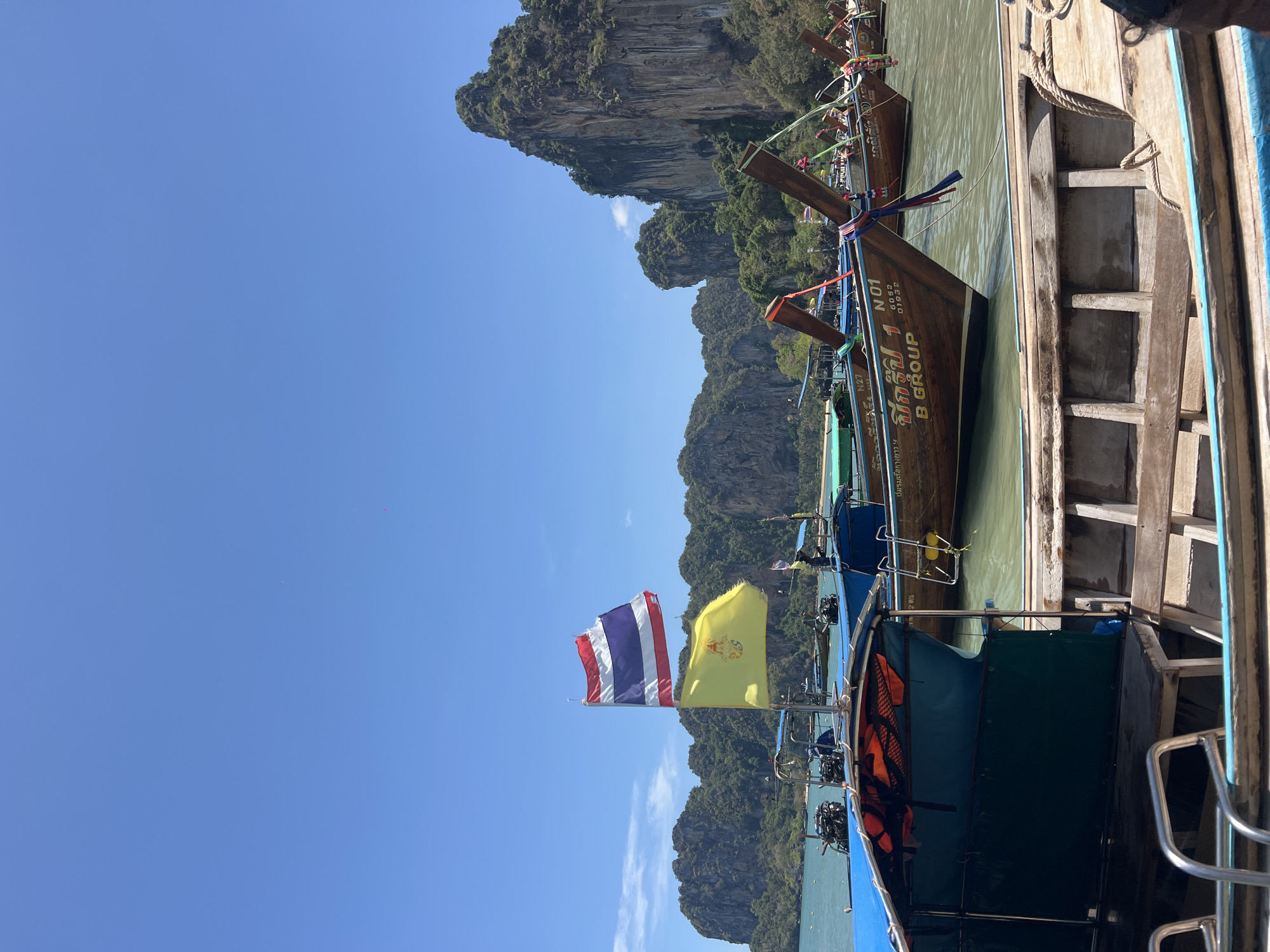 The image showcases several boats in Ban Ao Nang, Thailand. The boats are adorned with Thai flags and yellow flags, and surrounded by water. The background shows rocky cliffs covered in greenery under a clear blue sky. The boats vary in construction, with some being traditional longtail boats and others appearing more modern.