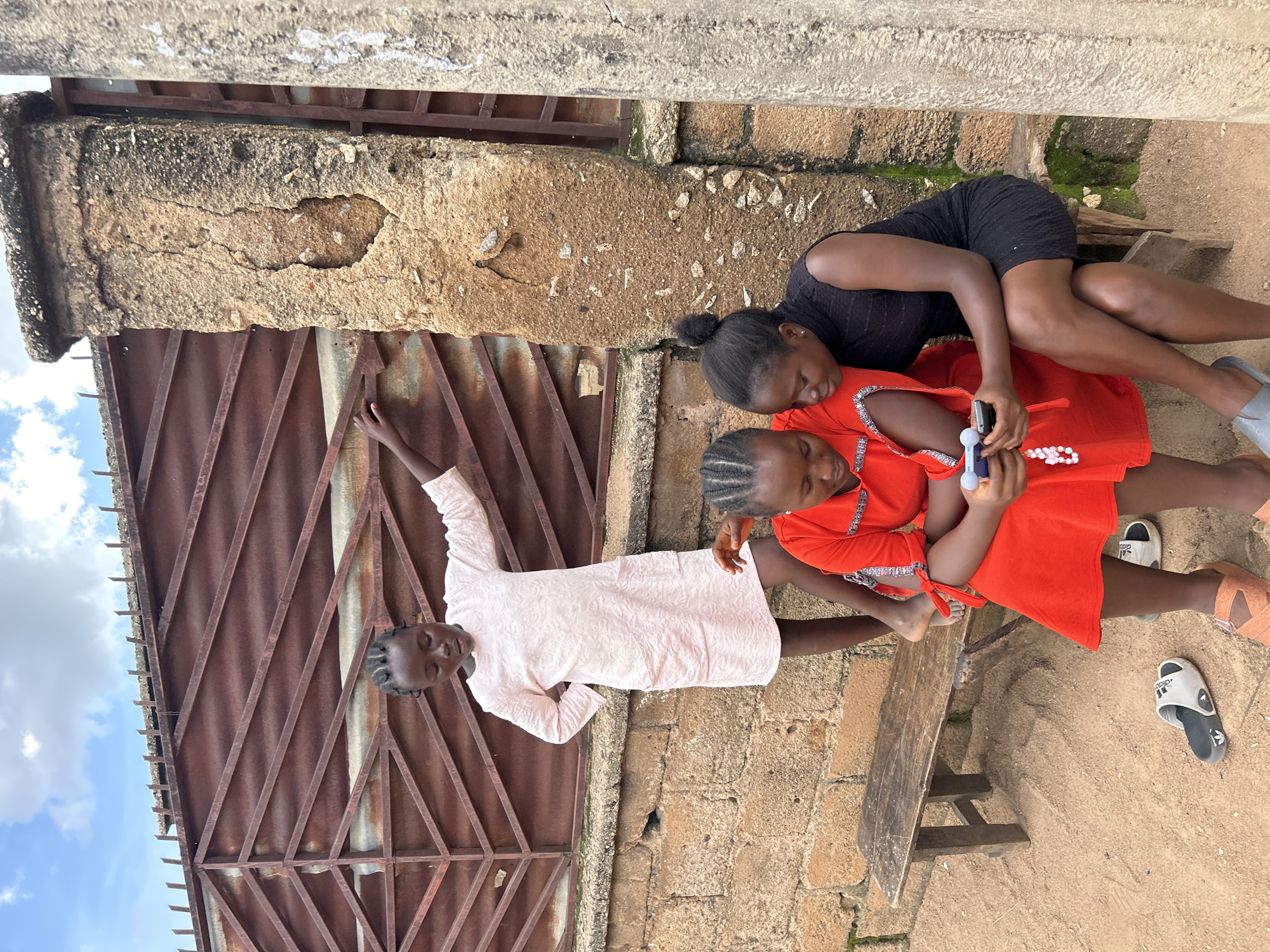 Three young Black females are in an outdoor setting. One girl, wearing a light pink dress, stands to the left, leaning against a rusty metal gate and a brick wall. Her left hand is on the gate, and her right hand is on her hip. Two other girls sit on a wooden bench. The girl on the left, wearing a bright red dress with white trim, holds a mobile phone in her hands. The girl on the right, wearing a black dress, looks at the phone with her head tilted. The ground is sandy, and there are a few pairs of sandals on the ground. A concrete pillar is on the right side of the image. The scene appears to be a residential area in Jos, Nigeria, characterized by its warm, dry climate and typical building materials.