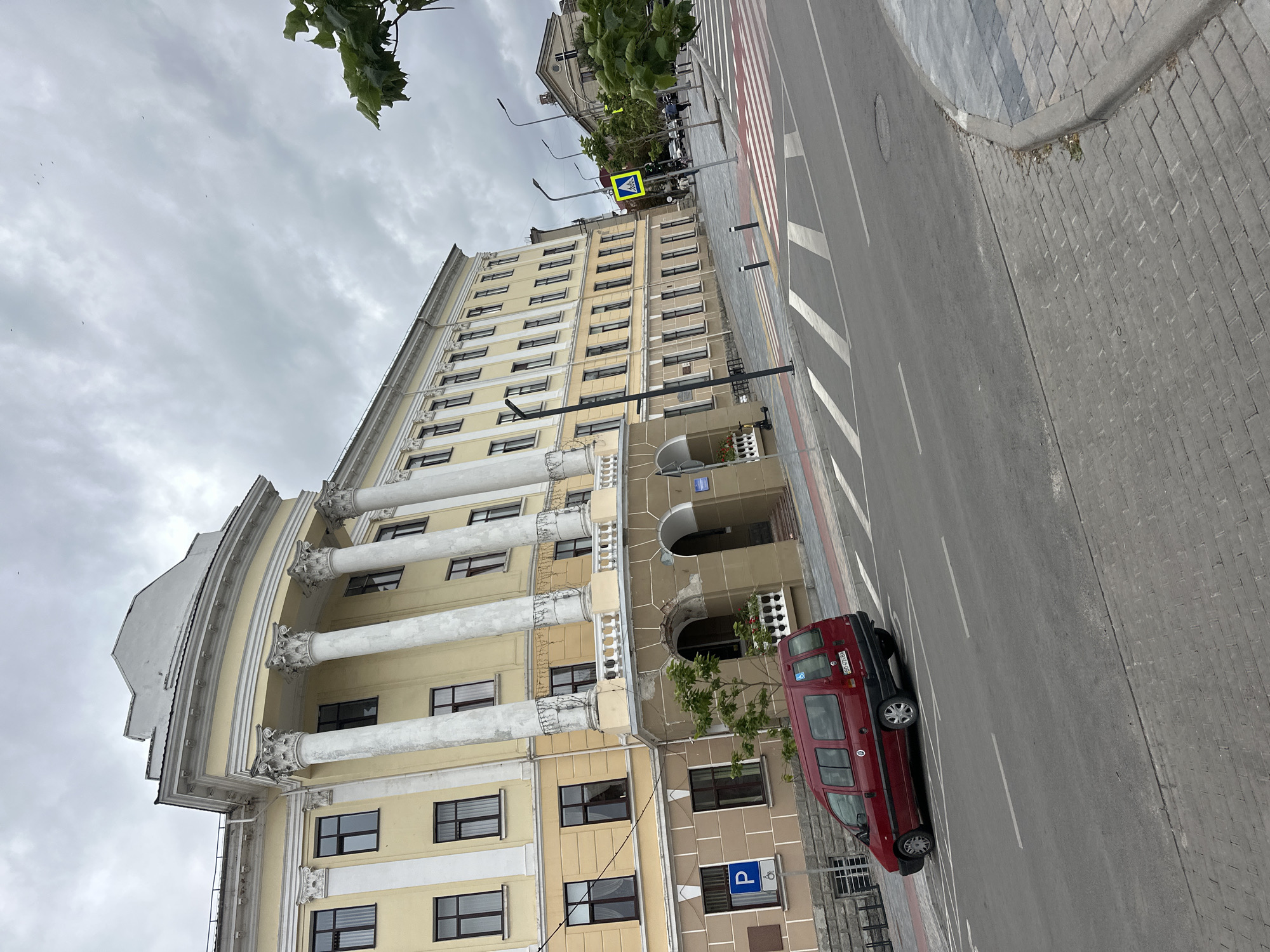 The image shows a light yellow building with classical architecture, featuring large white columns and a curved facade. In front of the building, a red minivan is parked near a designated disabled parking spot marked with a blue "P" sign. The street in front of the building has a pedestrian crossing with white stripes. The sky is overcast with grey clouds. The location is the Ternopil National Economic University in Ternopil, Ukraine, specifically the main building.
