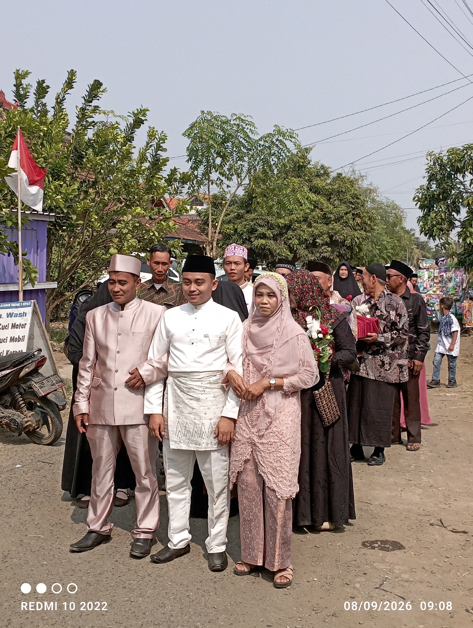The image shows a group of people dressed in traditional attire, possibly for a wedding or religious ceremony, standing on a dirt road in Dukuhseti, Indonesia. In the foreground, two men in matching suits and headwear stand next to each other, with the man on the right in a white outfit and the man on the left in a pale pink suit. Behind them, several other individuals, including men and women, are visible. One woman in the middle is holding a bouquet of flowers. The background features trees, houses, and a small stall with various items displayed. A motorcycle is parked on the left side of the image. The sky is clear and overcast. The timestamp "REDMI 10 2022" and "08/09/2026 09:08" are visible at the bottom of the image.