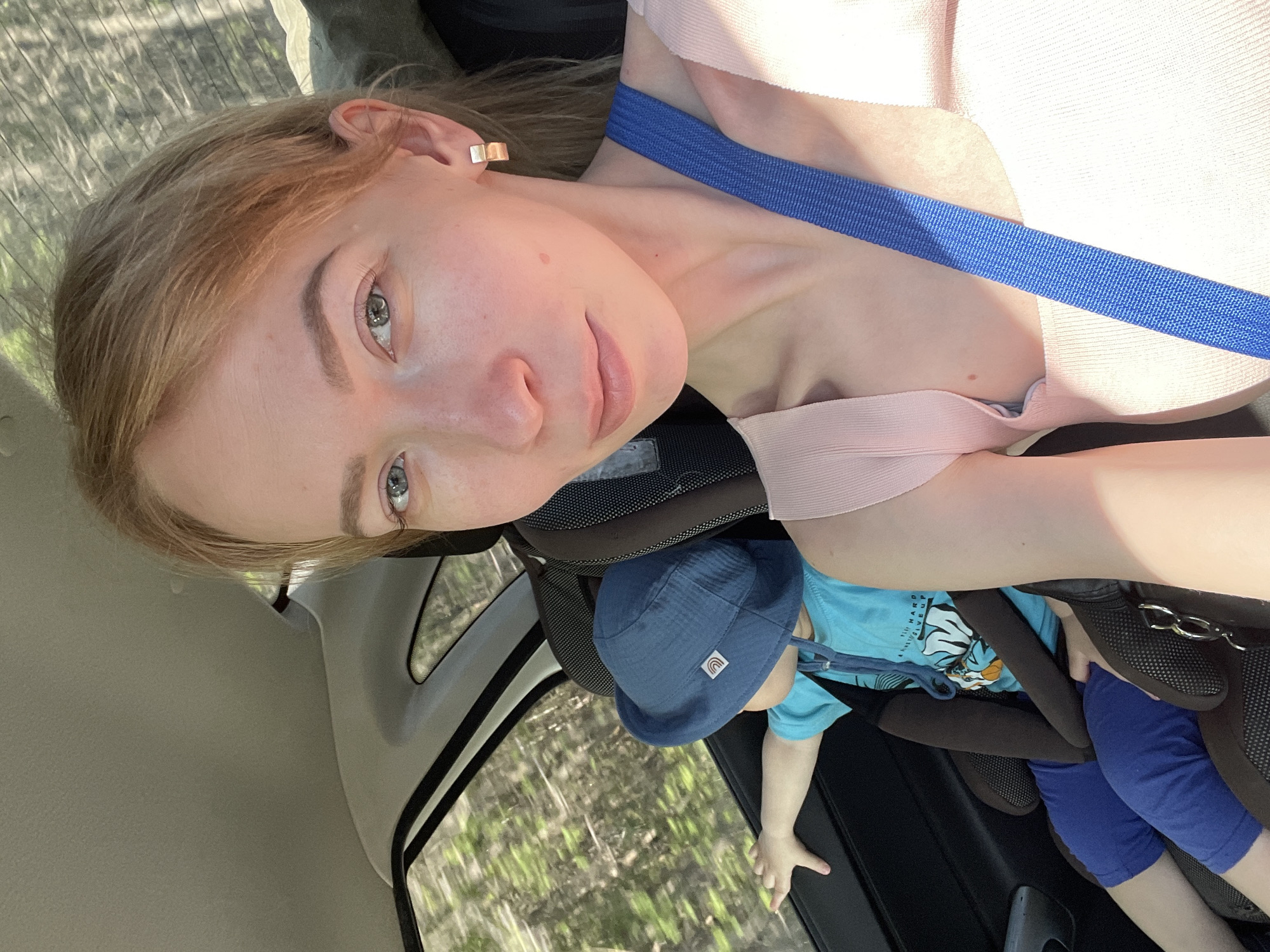 A fair-skinned woman with light brown hair is in the front passenger seat of a car. She has blue eyes, pink lips, and wears a square gold earring. A blue seatbelt crosses diagonally over her pink top. To her right, in the backseat, a young child wearing a blue hat, a turquoise shirt with a graphic print, and blue pants sits in a car seat, reaching towards the window. Trees and vegetation are visible through the car window. Based on the information provided in the prompt, the location is Perm, Russia.