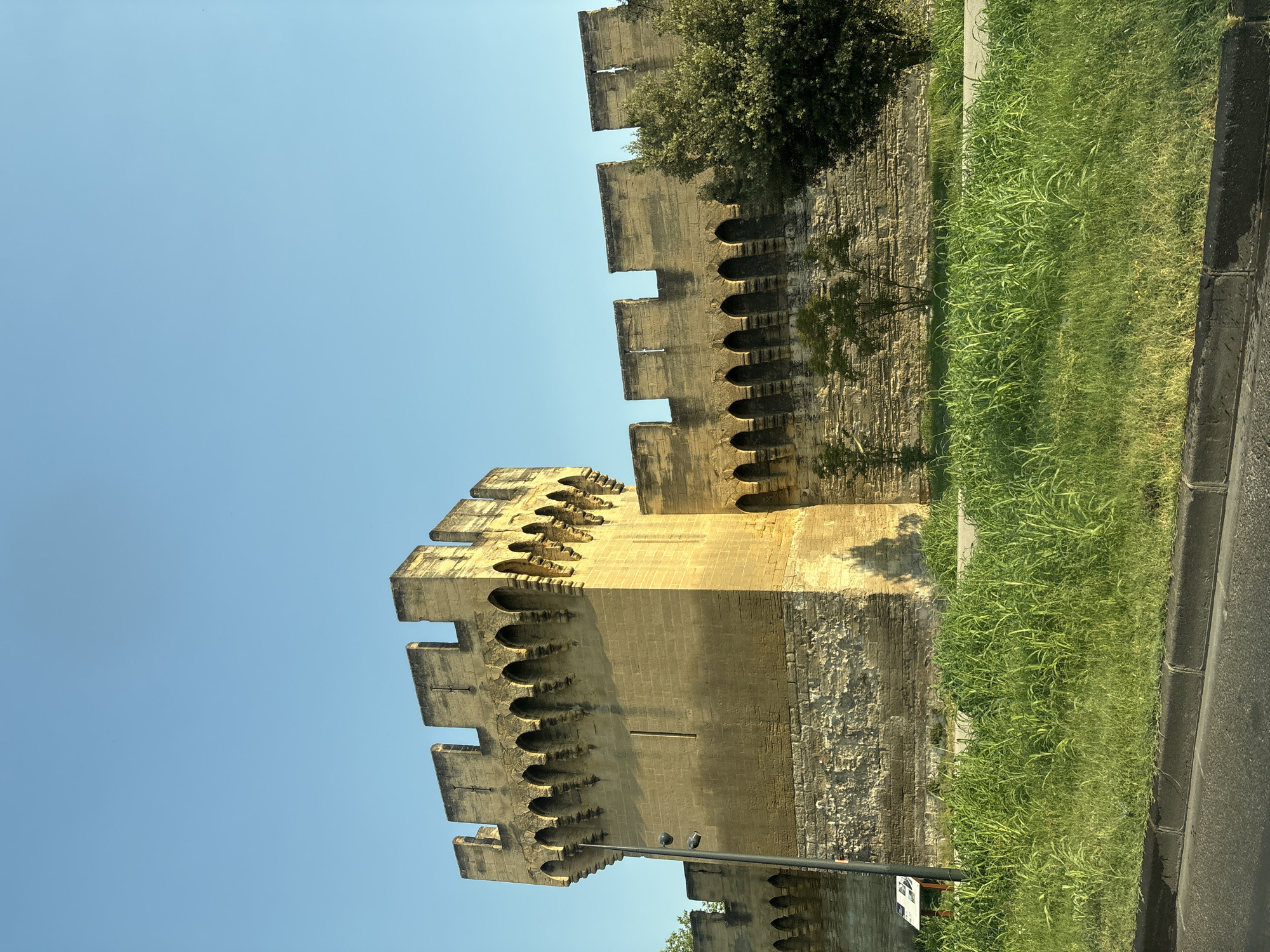 The image shows a section of the medieval city walls of Avignon, France, with a prominent tower on the left. The walls are constructed of large, light-colored stone blocks, weathered and showing signs of age. The top of the wall features crenellations, characteristic of fortifications. Below the crenellations, there is a decorative frieze with a series of arched openings. The tower is taller and more substantial than the wall, also made of stone blocks and topped with crenellations. A few small trees and a larger, dense green tree are visible behind the wall on the right side of the image. In the foreground, there is a strip of lush green grass and a paved area, likely a sidewalk, with a dark curb and asphalt road in the very bottom. The sky is a clear, bright blue, suggesting a sunny day. There are no people visible in the image.