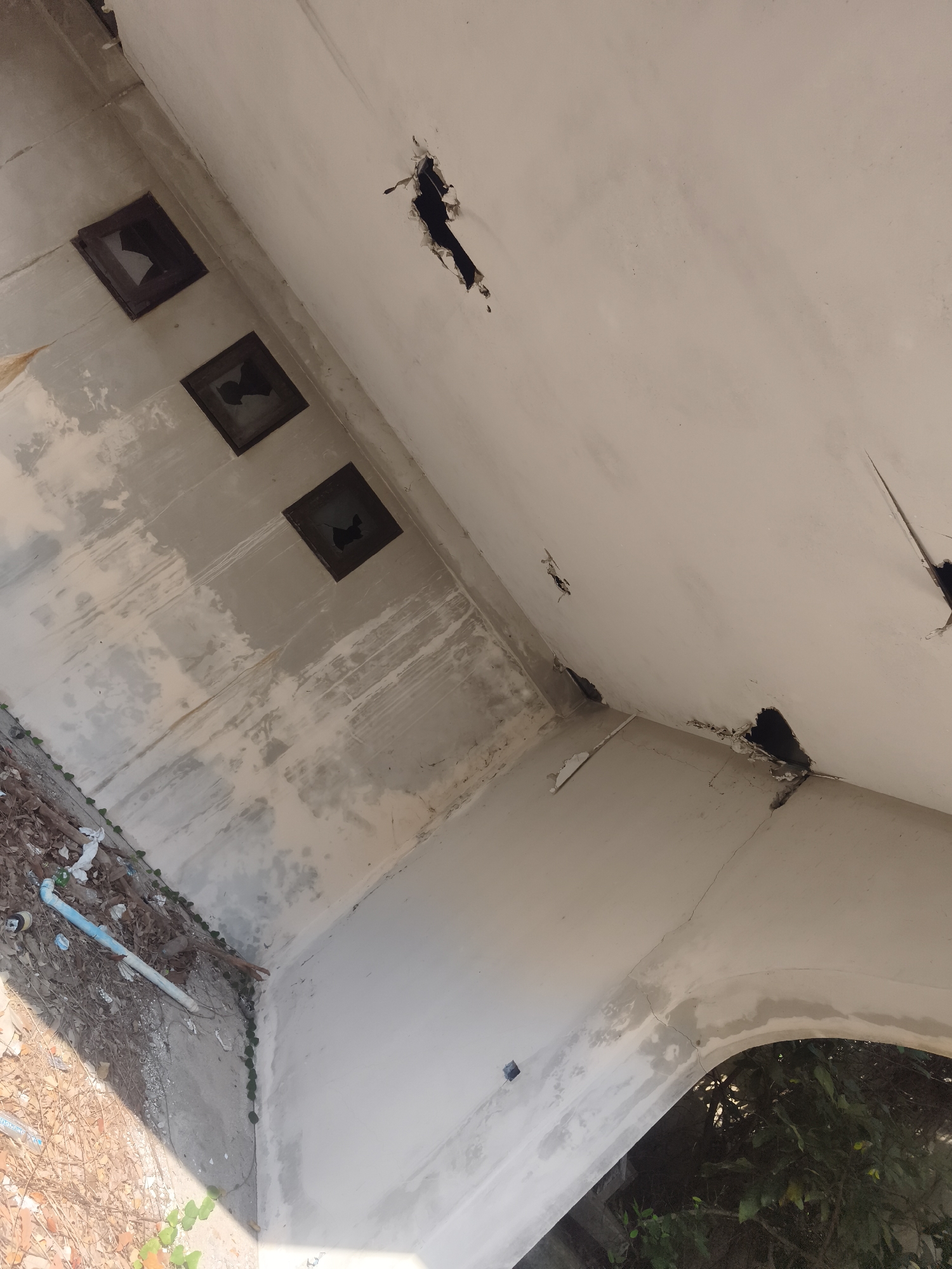 The image shows the interior of a derelict structure in บ้านบางรักษ์, Thailand. The walls are partially plastered and stained, with significant damage including large holes in the ceiling. Three square openings in the wall are visible, each containing broken fragments. Debris and vegetation surround the base of the walls. The perspective is angled upwards, focusing on the damaged structure.