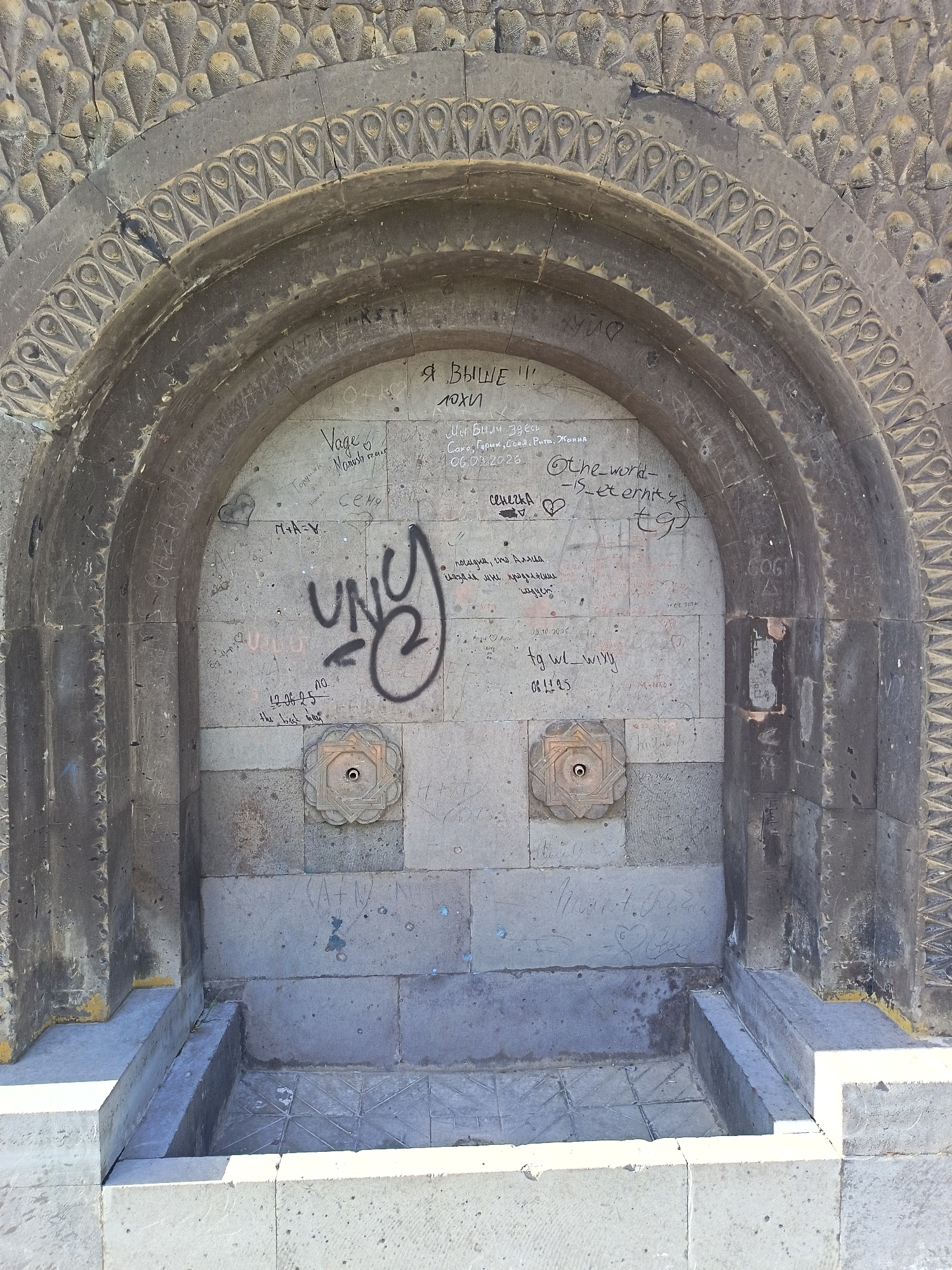 The image shows a weathered stone structure, likely a public water fountain or monument, covered in graffiti. The structure features an arched opening with ornate carvings around the edges and two decorative spouts on the back wall. The graffiti consists of various names, dates, phrases in multiple languages, and tags, including the large tag "UNC". The fountain's basin appears empty and constructed from the same stone as the rest of the structure. The location is identified by the tags as in city of Unknown, Unknown.