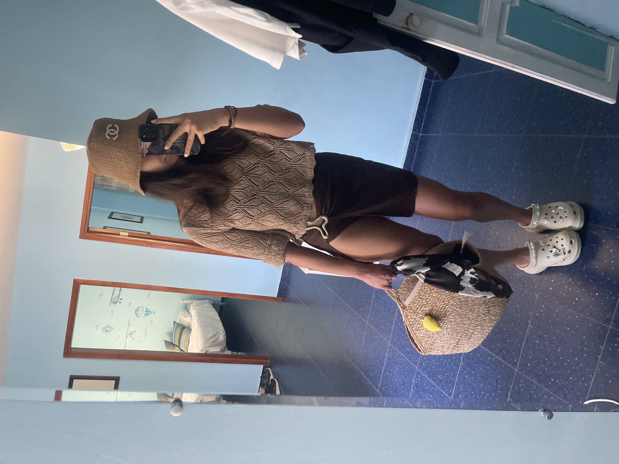 A young woman stands in a room with blue walls and a tiled floor. She is wearing a knitted cropped top, a brown wrap skirt, a woven bucket hat with a Chanel logo, and decorated Crocs. She holds a woven bag with a black and white scarf and a yellow accessory. She is taking a mirror selfie with her phone. In the background, through an open doorway, a bedroom with a bed and hot air balloon decals on the wall is visible. Another doorway reflects a similar bedroom. Clothes are hung on a rack to the right of the frame. The scene appears to be in a residential setting in Chernomorets, Bulgaria.