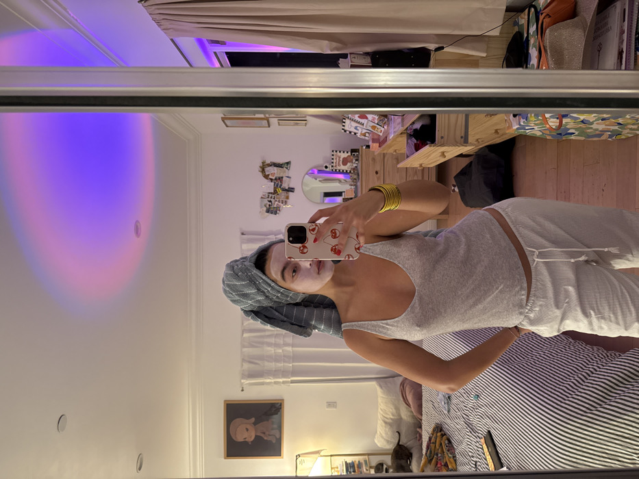 The image depicts a female subject taking a mirror selfie in a residential bedroom in Los Angeles, California, United States of America.
The subject is a young adult female, standing upright, with her hair wrapped in a grey and white striped towel. Her face is covered with a white facial mask. She is wearing a light grey V-neck tank top and white and light grey striped pajama bottoms secured with a white drawstring. Her left hand is placed on her hip, and her right hand holds a white smartphone with a red cherry-patterned case, pointed towards the mirror to capture the reflection. Multiple yellow bangles are visible on her right wrist.
The bedroom features a white ceiling with recessed lighting and a circular projection of purple and pink gradient light. To the left, a bed with a black and white striped duvet and white pillows is partially visible, along with a small black cat resting on a pillow. A floor lamp with a white shade and a framed artwork, depicting a cartoonish sad girl with dark hair, are positioned to the left of the bed. To the right, a light-colored wooden dresser with multiple drawers, some ajar, is visible. Various decorative items, including a small mirror and framed pictures, are arranged on the dresser and the wall above it. A purple LED light strip is visible behind light-colored curtains on the far right, reflecting in the mirror. The floor is covered with light-toned wood.