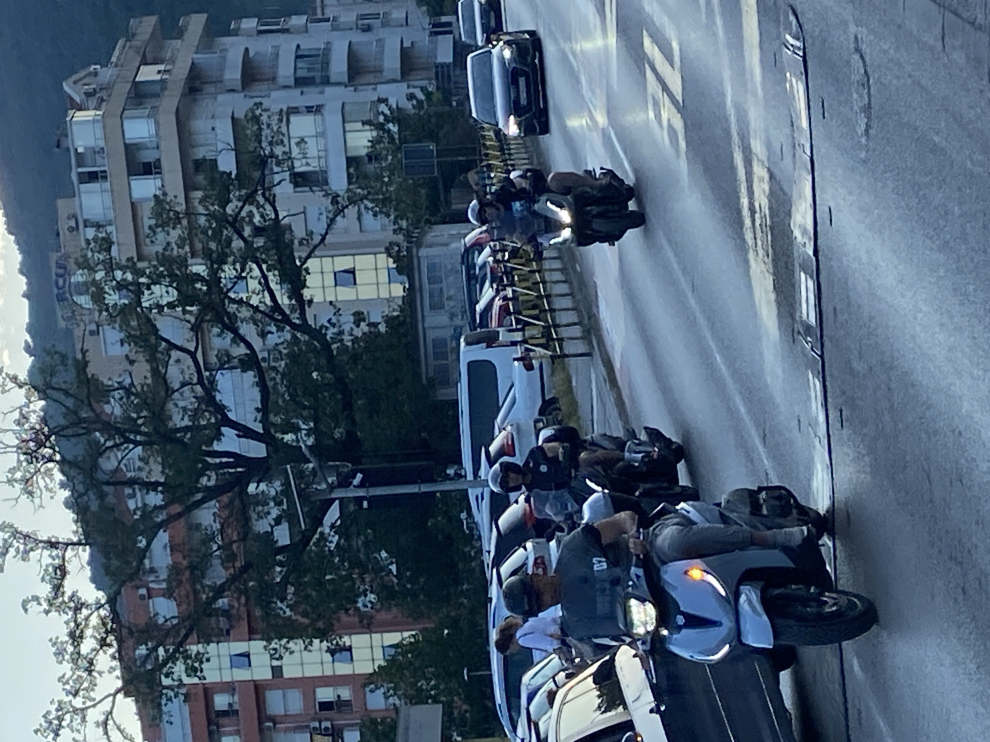 The image depicts a street scene in Budva, Montenegro, likely during daylight with overcast conditions, possibly early morning or late afternoon given the lighting. In the foreground, a person is riding a light-colored scooter, wearing a dark t-shirt and grey pants, and a dark helmet. The scooter has its headlights and an orange turn signal illuminated. To the right, another scooter is visible with a rider wearing a dark helmet and a dark uniform-like shirt, riding in the same direction. Behind these scooters, a dark-colored car is moving in the right lane. Several other vehicles, including cars and a white van, are present further in the background, suggesting moderate traffic. The road surface shows speed bumps and lane markings, including a faded "50" indicating the speed limit. Flanking the road are modern apartment buildings with multiple balconies and large windows, reflecting the surrounding environment. Trees with dense foliage are interspersed, some obscuring parts of the buildings. The overall impression is a typical urban street in a coastal city.