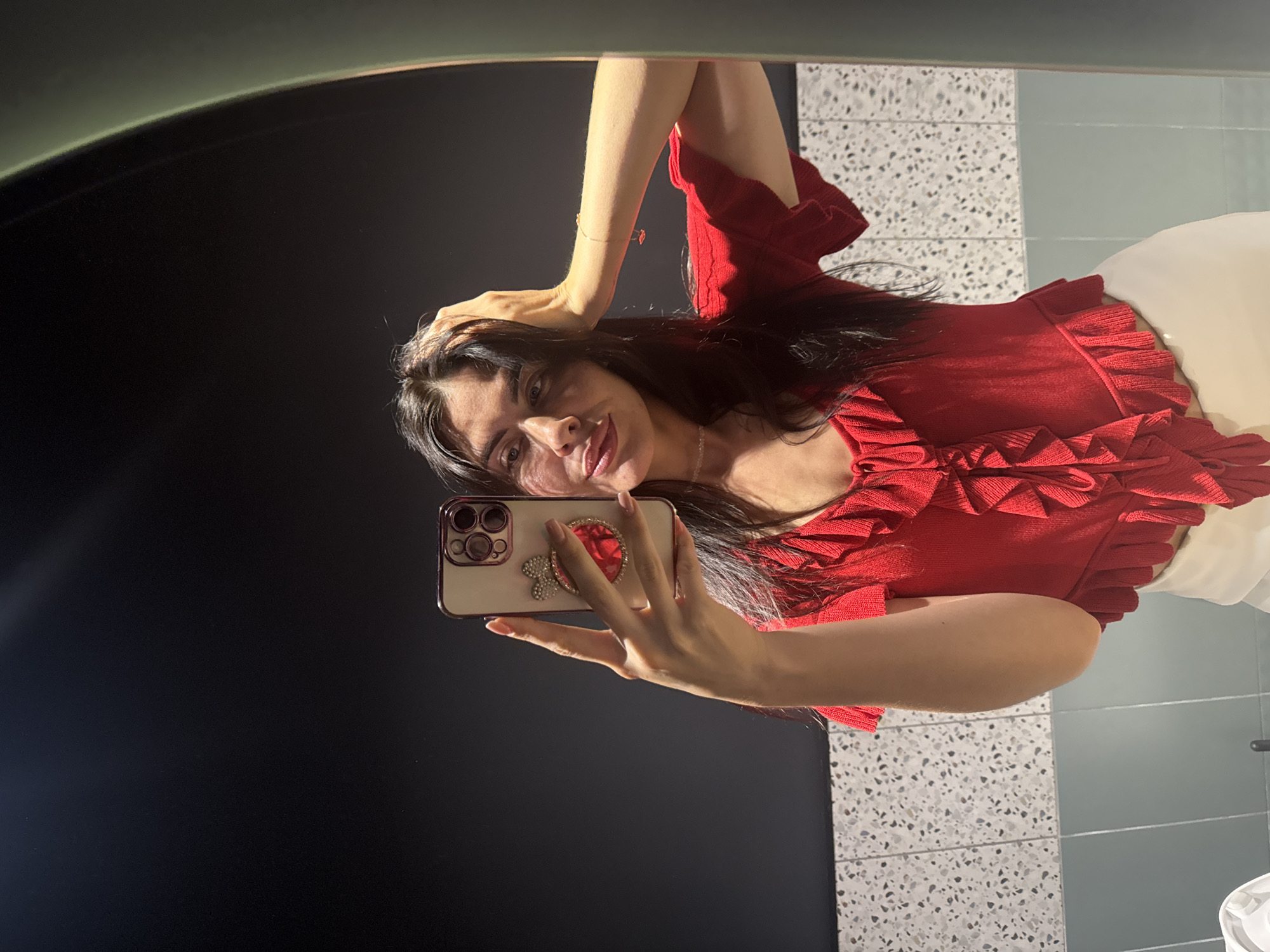 A young woman with long, dark hair is taking a mirror selfie in a dimly lit bathroom. She is wearing a red, ruffled top and white pants. Her right hand is raised, holding a smartphone adorned with a pink bow and a red jewel. Her left hand is resting on her head. The background shows a dark wall, a speckled tiled wall, and a light green-tiled wall, reflecting in a large, curved mirror.