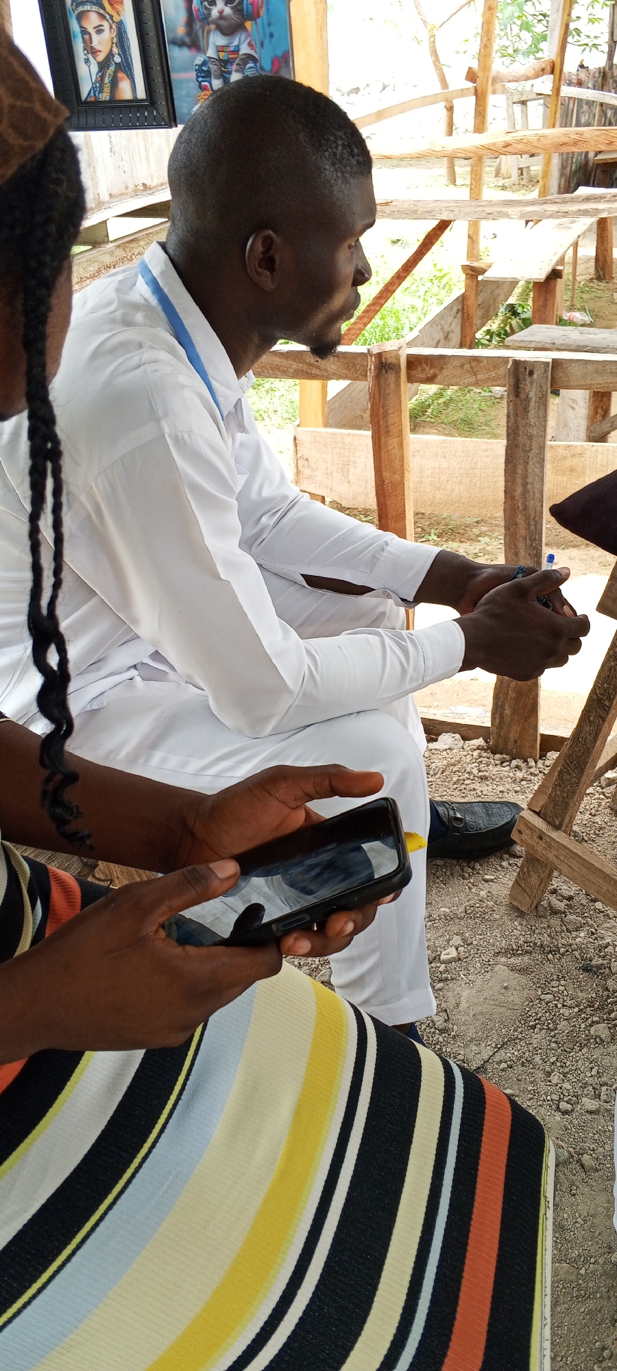 The image depicts a scene in what appears to be an outdoor setting, possibly in Keffi, Nigeria. Two individuals are visible; a man in a white shirt and pants sits with his hands clasped. The second individual, closer to the camera, is using a smartphone. She is wearing a striped shirt. The background includes what appears to be a wooden structure, possibly a bench or platform. Also, two framed pictures are attached to a wooden post behind the man wearing white; one is of a woman, and the other is of a cat wearing a t-shirt and headphones. The ground is dirt or gravel.