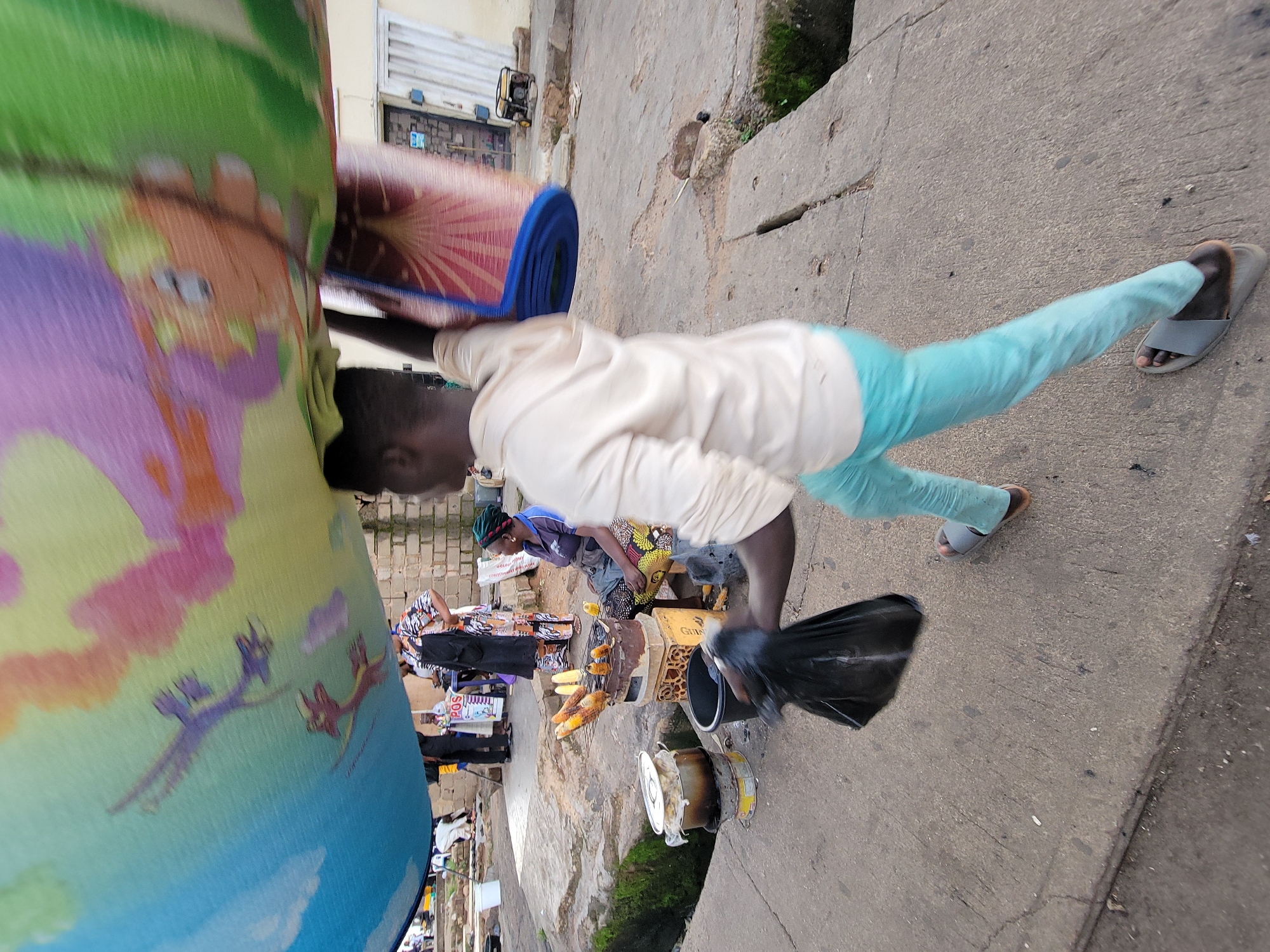 A person wearing a light-colored hoodie and light blue pants walks on a concrete surface, carrying a rolled-up mat on their head and a black plastic bag in their right hand. The mat is large and colorful, with a pattern of what appear to be purple dragons or similar creatures against a blue and yellow background. To the left of the walking person, a woman sits on a low stool, wearing a headscarf and a patterned dress, attending to a grill where several ears of corn are being roasted. In the background, other individuals are visible, along with market stalls and brick buildings. The scene appears to be a street market in Jos, Nigeria.