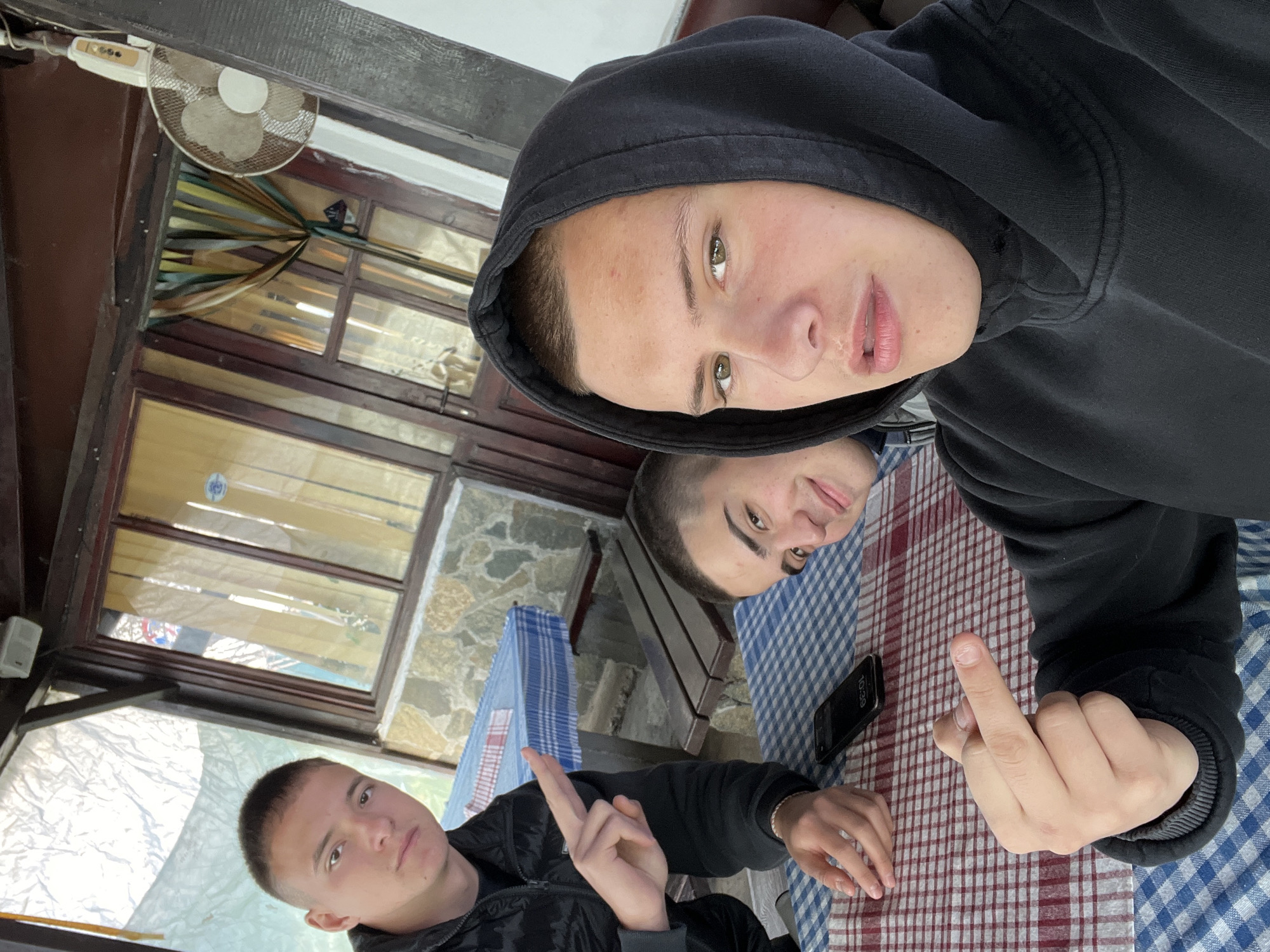 Three young males are visible in an outdoor setting. The individual in the foreground is wearing a black hooded sweatshirt and is extending their middle finger. To their right is a smartphone on a checkered tablecloth. Behind them, two additional males are visible; one is seated and also extending their middle finger, and another is looking at the camera. The location appears to be a covered terrace or patio. In the background, windows and stone walls are visible. A fan is seen mounted on the ceiling in the upper left corner. It is not possible to determine the precise location in Asenovgrad, Bulgaria, without additional contextual information.
