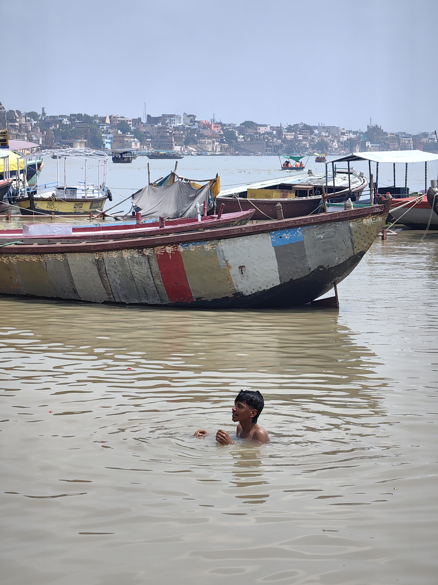 A young boy is swimming in the Ganges River in Varanasi, India. He is partially submerged in the murky water, with his head and shoulders visible. His hands are clasped in front of him, and he is looking up and to his right. In the background, several boats are moored along the riverbank. The boats are of various sizes and colors, some with awnings and others empty. Beyond the boats, a cityscape of buildings, temples, and ghats stretches along the horizon. The sky is overcast, and the overall atmosphere is hazy. The water is a muddy brown, with ripples and reflections from the boats and the sky.