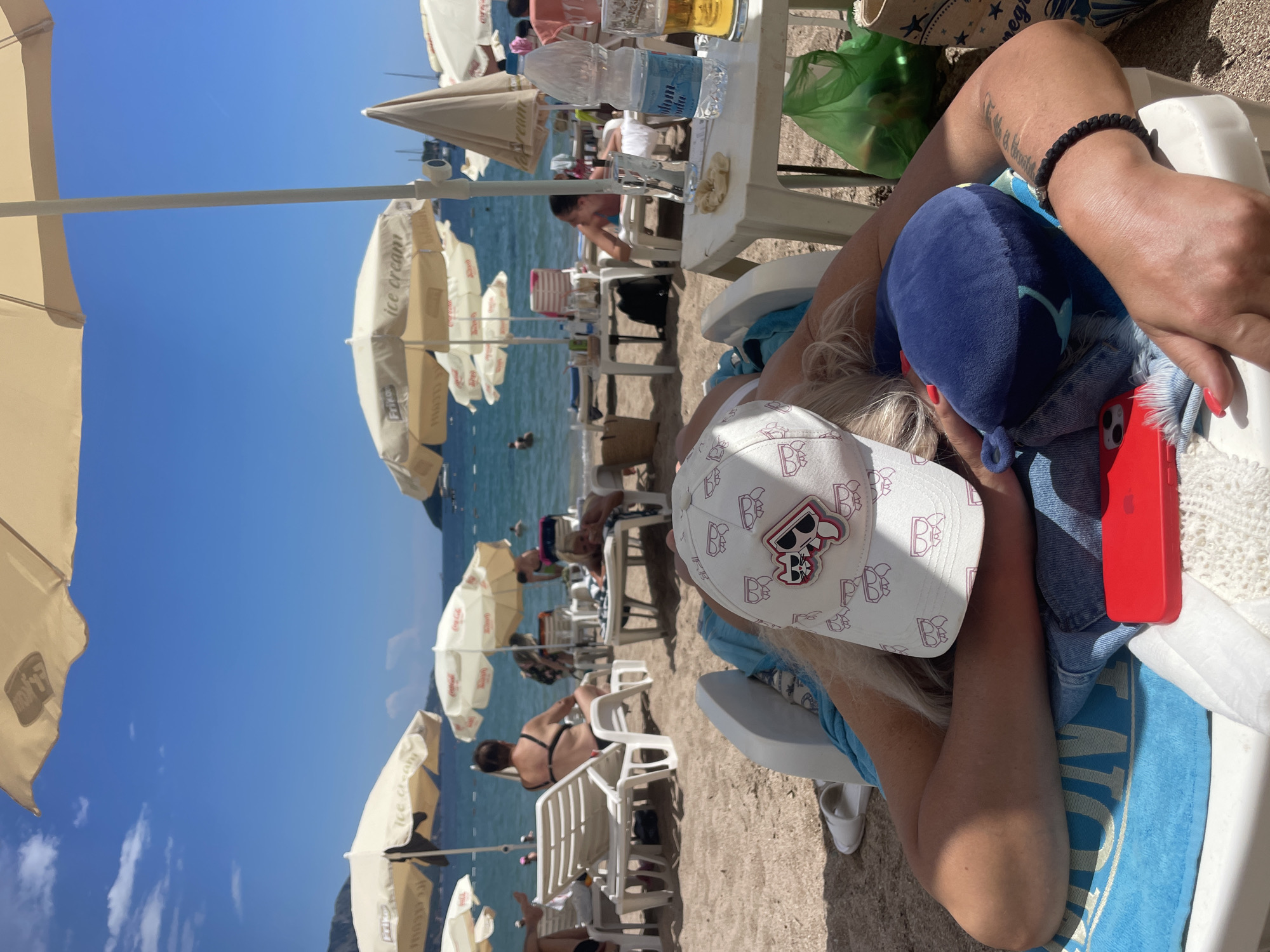 This image depicts a sunny day at a beach in Budva, Montenegro. The foreground features a woman reclining on a white lounge chair, partially obscured by a blue plush toy and a white baseball cap adorned with a cartoon skull logo. Her arm is visible, wearing a black beaded bracelet. A red iPhone is placed beside her.
The background reveals a bustling beach scene. Numerous white and beige umbrellas are set up on the sand, providing shade. Several beach chairs are occupied by people. The sea is calm and blue, with a few individuals visible in the water. In the distance, hills or mountains are faintly visible.
The overall atmosphere suggests a relaxed vacation setting. The bright sunlight indicates it's daytime, likely midday or early afternoon. Text visible on some of the umbrellas includes "ice cream" and "Frikol." The presence of lounge chairs, umbrellas, and people enjoying the beach suggests a tourist-oriented location.