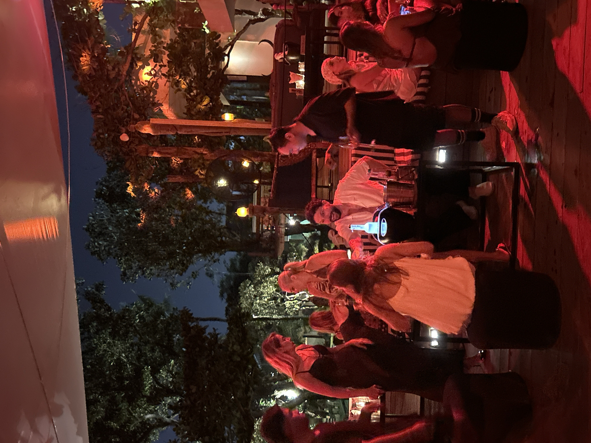 The image depicts an outdoor evening gathering at a venue in North Kuta, Indonesia, possibly a bar or restaurant. Red ambient lighting dominates the scene, casting a warm glow on the patrons and surroundings. Several individuals are visible, engaged in conversation and socializing. In the foreground, a woman in a black dress stands near a table, while another woman in a white top is seated on a dark cylindrical stool. To the right, a man in a black shirt and shorts stands and appears to be interacting with a group of people. Behind them, more individuals are gathered around tables, with trees and foliage forming the background. The overall atmosphere suggests a casual and lively social event.