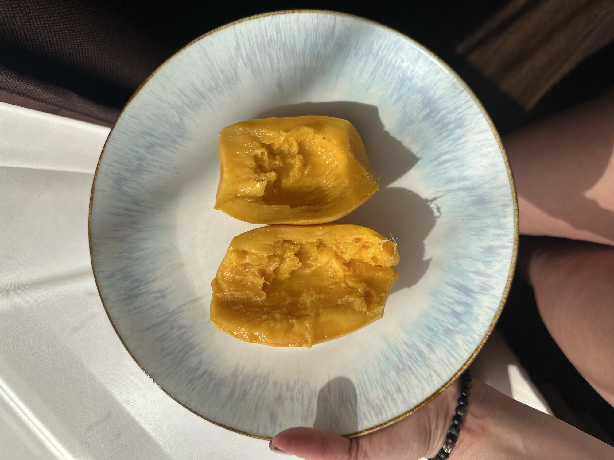A top-down, close-up shot shows two pieces of carved ripe mango on a round white plate with blue streaks, held by a person. The plate's edge is trimmed with a thin brown line. The mango pieces are irregularly shaped and deeply yellow, with textured surfaces suggesting they have been cut or scooped from the fruit. The person holding the plate is partially visible, with a hand wearing a beaded bracelet and a section of their leg in the background. The lighting appears to be natural, with distinct shadows cast by the mango pieces on the plate. The background is varied, consisting of dark brown and white areas that appear to be furniture and clothing. The location is unknown, and there is no way to determine if this is a photo of Chaikovsky, Russia.