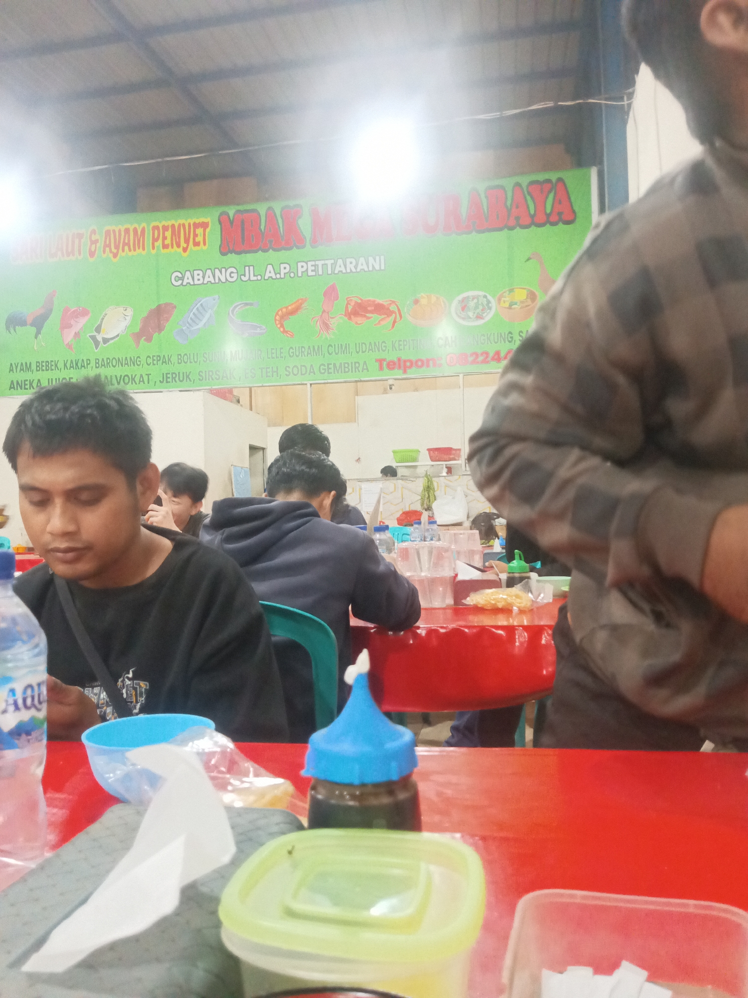 The image depicts an indoor dining area, possibly a restaurant, with multiple individuals present. In the foreground, a man with dark hair and a black t-shirt is seated at a red table, looking down. To his right, a person wearing a camouflage patterned shirt is partially visible, also seated at the table. Several plastic cups and containers are arranged on the table, along with a bottle of water. Behind the man in the black t-shirt, other patrons are seated, some with their backs to the camera. A large green banner with text in Indonesian is visible in the background. The banner appears to advertise seafood and chicken dishes, with the words "SURABAYA" and "CABANG JL. A.P. PETTARANI" prominently displayed, indicating the location is a branch on Jalan A.P. Pettarani in Surabaya, but the user states the location is Makassar, Indonesia. The environment seems casual and crowded, suggesting a busy dining establishment.