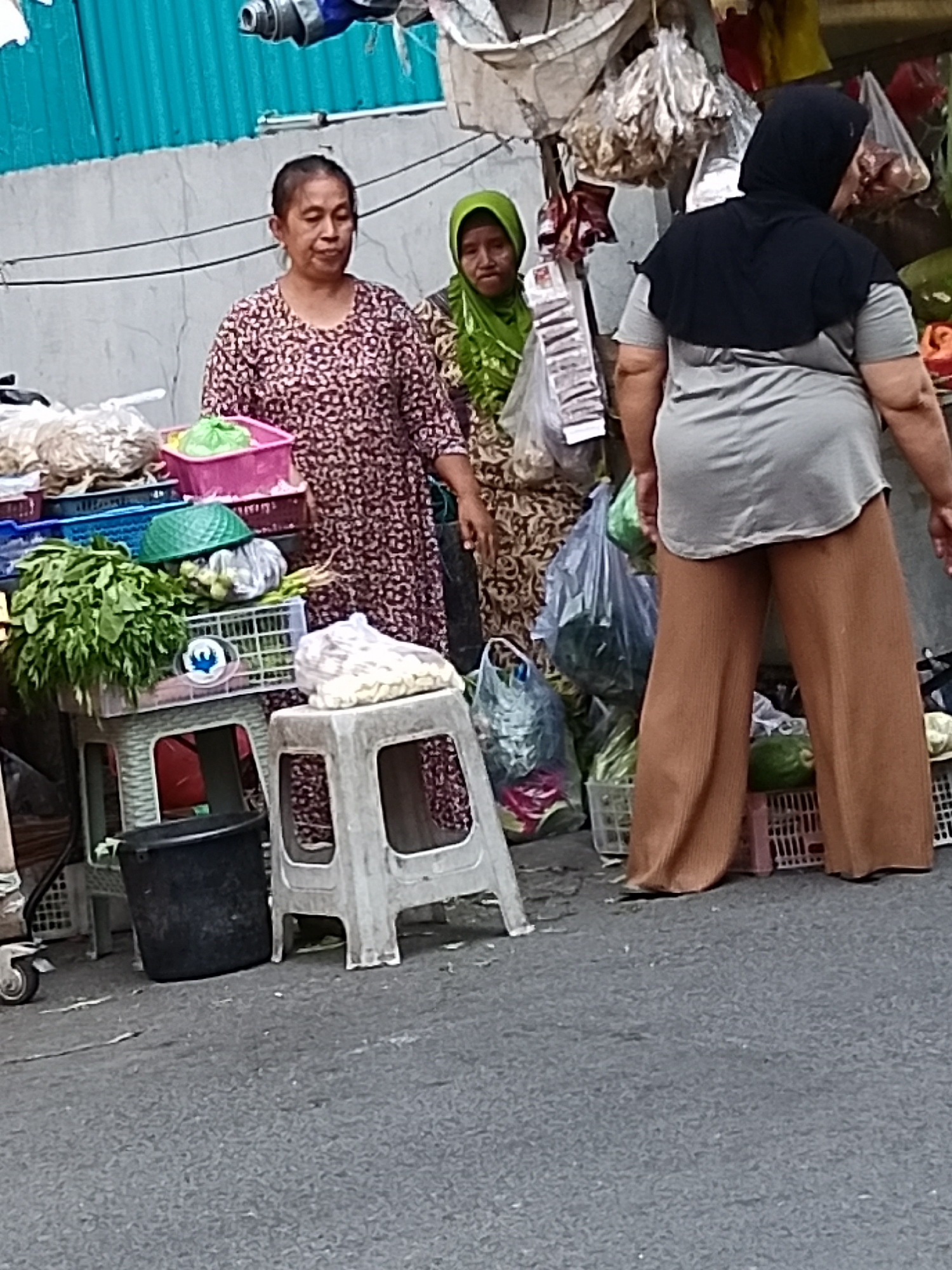The image depicts a street scene in Surabaya, Indonesia, possibly a market stall. Three individuals are present. On the left, a woman with short, dark hair wears a patterned dress. In the center, a woman with a green hijab and a patterned top stands partially obscured. To the right, a person wearing a black hijab and a grey t-shirt with brown wide-leg pants faces away from the camera. The background shows various goods, including vegetables, possibly dried fish hanging, and stacked crates. The ground is paved with asphalt, and debris is scattered. The overall setting suggests an outdoor market or roadside vending area, typical of urban environments in Surabaya.