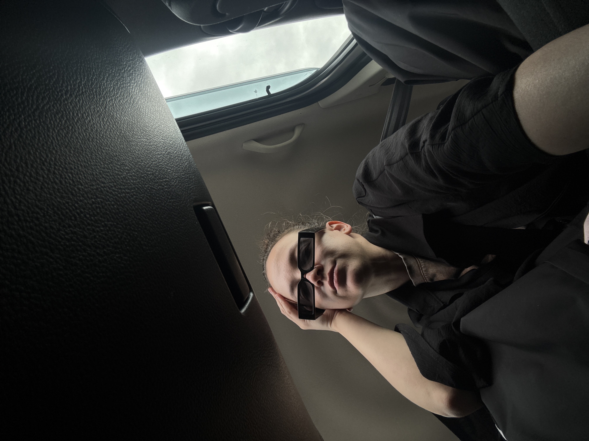 A person with fair skin and curly hair is lying down in a car. They are wearing black clothing and blocky black sunglasses that cover the area between their eyes and upper lip. Their left hand is positioned with the palm facing upwards and their face resting on their hand. The camera angle is looking upwards from below the person's face. The car's interior is visible, with the door panel on the left, showing black textured material. A sunroof is partially open above the person, revealing a cloudy sky. The overall lighting is dim and somewhat stark. There is no evidence to suggest that the location is Bol'shoi Mashlyak, Russia.