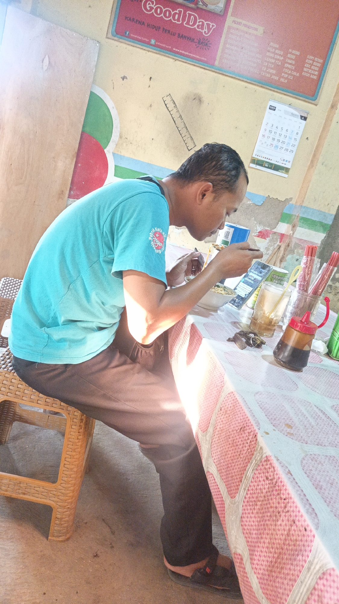 A person is seen sitting at a table in what appears to be a roadside eatery in Dawe, Indonesia, consuming a meal and looking at a mobile phone. The person is wearing a turquoise t-shirt and black pants, and is seated on a brown woven plastic chair. On the table, there is a bowl of noodles, a glass of iced tea, a bottle of soy sauce, and a container of chopsticks. The background shows a yellow wall with some colorful markings and a calendar indicating August 2014. The scene is illuminated by bright sunlight, casting strong shadows.