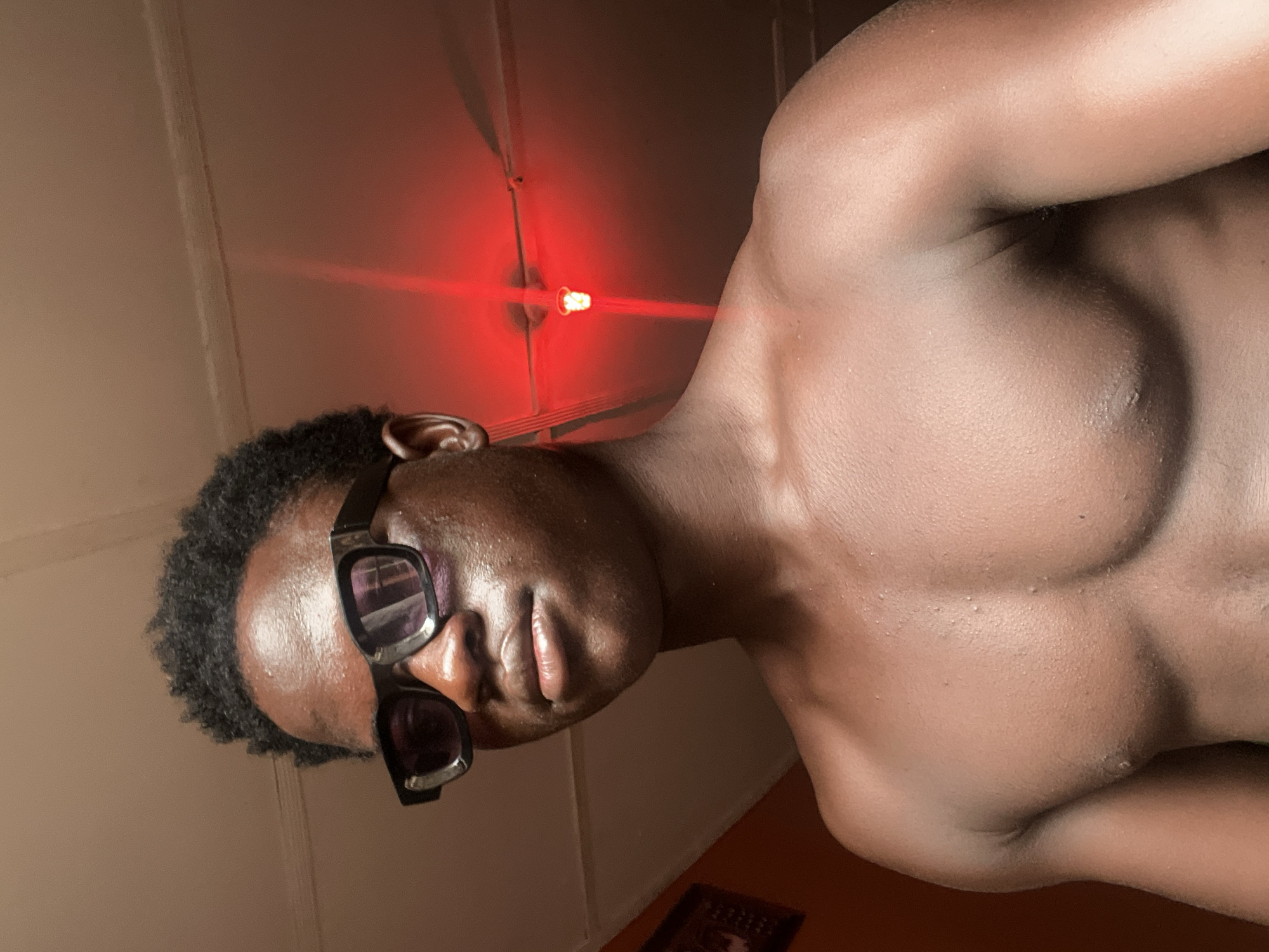 A close-up, low-angle indoor photograph shows a shirtless dark-skinned man wearing black sunglasses. He has short, dark curly hair. His upper torso and shoulder are visible. Above him is a ceiling with a light fixture, emitting a red glow. A small rectangular object is partially visible in the bottom portion of the image. The location is unknown, and there is no indication of it being in the city of Mosafejaw, Nigeria.