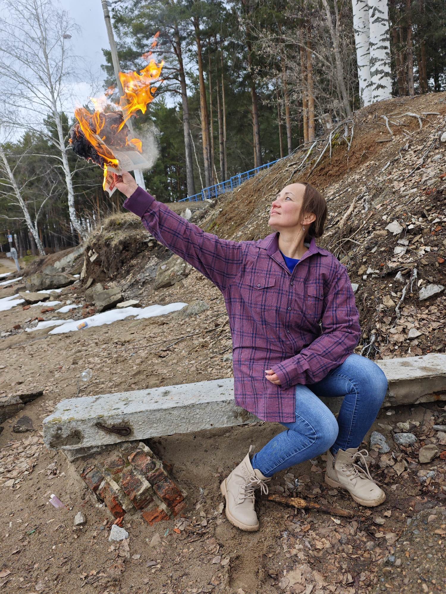 A woman sits on a concrete block amidst an eroded landscape with birch and pine trees, holding a burning object aloft. The location is likely the city of Chaikovsky, Russia, based on visual cues in the background and the prevalence of birch trees, but I am unable to ascertain any specific details about the location from the image. The woman is wearing jeans, and a purple button-down flannel shirt. The ground is covered with loose dirt, fallen leaves, bits of brick, and some remaining patches of snow. The fire appears to be consuming a mass of paper, or some other organic material, which is creating smoke. The sky is overcast.