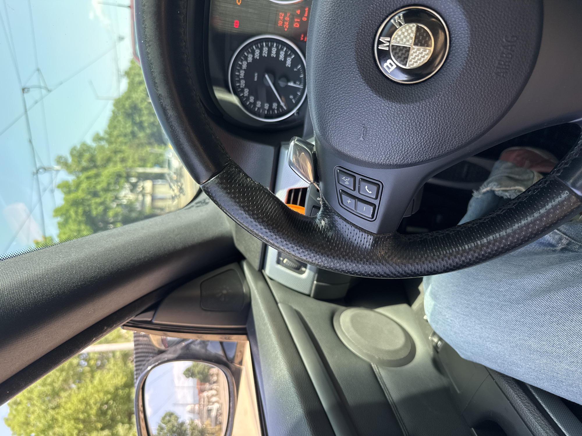 The image shows the interior of a BMW car, focusing on the steering wheel, dashboard, and side mirror. The steering wheel has the BMW logo and integrated controls. The speedometer displays a range from 0 to 280 km/h. A side mirror is visible, reflecting an urban environment with buildings and trees. A person wearing blue jeans is in the driver's seat. The time on the dashboard indicates 10:42, with a temperature reading of 24.0 degrees Celsius.