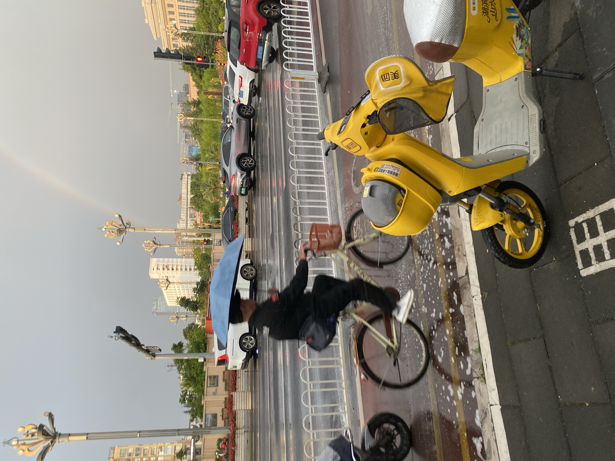 In Xishan District, China, on a rainy day, a person rides a bicycle under a blue umbrella across a wet street. The rider is wearing dark clothing and white sneakers. In the foreground, a bright yellow electric scooter is parked. Behind the cyclist, cars are stopped at an intersection, and a subtle rainbow is visible in the overcast sky. Tall buildings and streetlights line the background. The scene is one of urban activity persisting despite inclement weather.