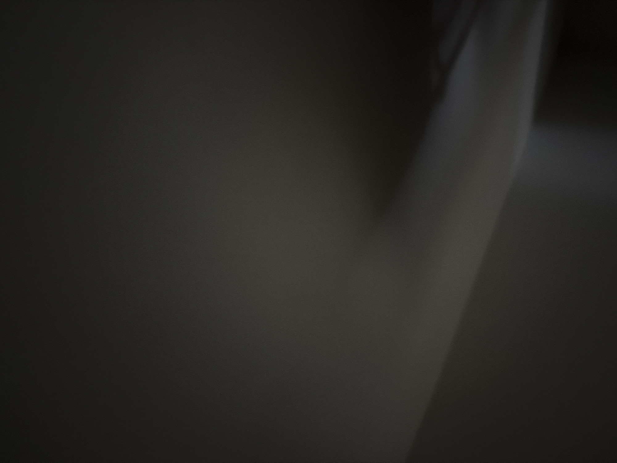 The image is a very dark, blurry, and abstract shot of a room or hallway. A diagonal line of lighter color, possibly indicating a wall or a curved surface, cuts through the left side of the frame. The rest of the image is uniformly dark gray and appears to have a grainy texture. No specific location in The Hague, Netherlands, can be identified from this image. There are no discernible people, subjects, objects, actions, or interactions.