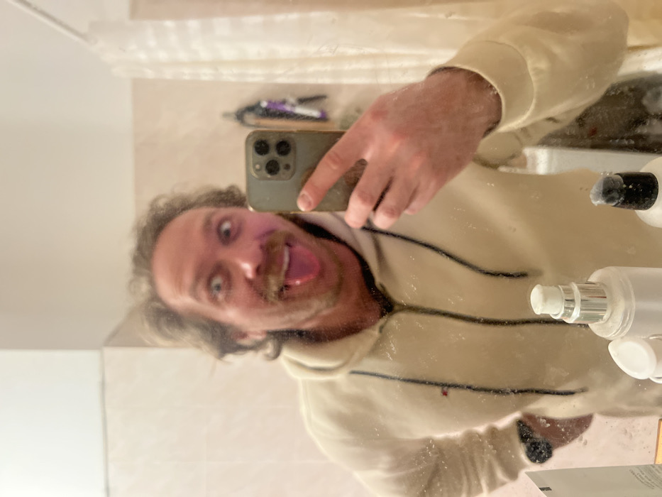 A male subject is depicted in a mirror selfie taken within what appears to be a bathroom environment in Saint Petersburg, Russia. The subject, exhibiting a humorous expression with wide eyes and an extended tongue, possesses light brown, wavy hair and a mustache. He is attired in an off-white hooded sweatshirt featuring a dark zipper, dark drawstrings, and a small multi-colored emblem on the left chest. A black wristwatch is present on his left wrist. The subject holds a dark-colored smartphone, equipped with a triple-lens camera array, in his left hand, positioned to photograph his reflection. The mirror surface displays numerous smudges or water droplets. Situated on a counter beneath the mirror are several toiletry bottles, including clear bottles with silver and black pump dispensers. The reflected background reveals a light-toned wall, a metallic shower curtain rod supporting a white curtain, and a wall-mounted hook rack with hanging items.