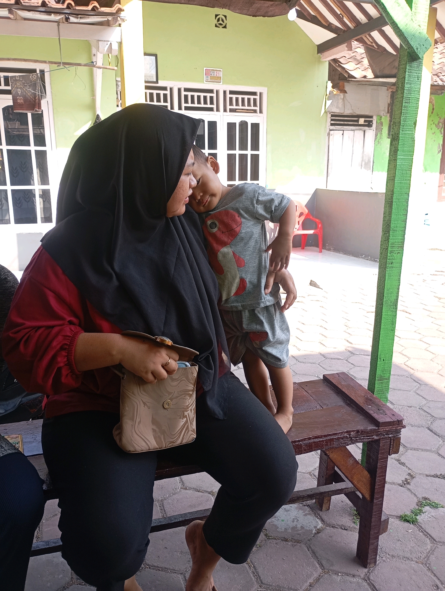 A Muslim woman wearing a black hijab and a maroon long-sleeved shirt embraces a young boy wearing a grey t-shirt and shorts with a red and white cartoon character print. The woman is seated on a wooden bench, holding a small tan purse with a marble pattern. The boy is standing, leaning into the woman. They are outdoors in what appears to be a residential area in Babelan, Indonesia. In the background, a light green painted building with white window frames and a tiled roof is visible, along with a wooden pillar painted green. The ground is paved with hexagonal stone tiles. Sunlight casts shadows on the ground.