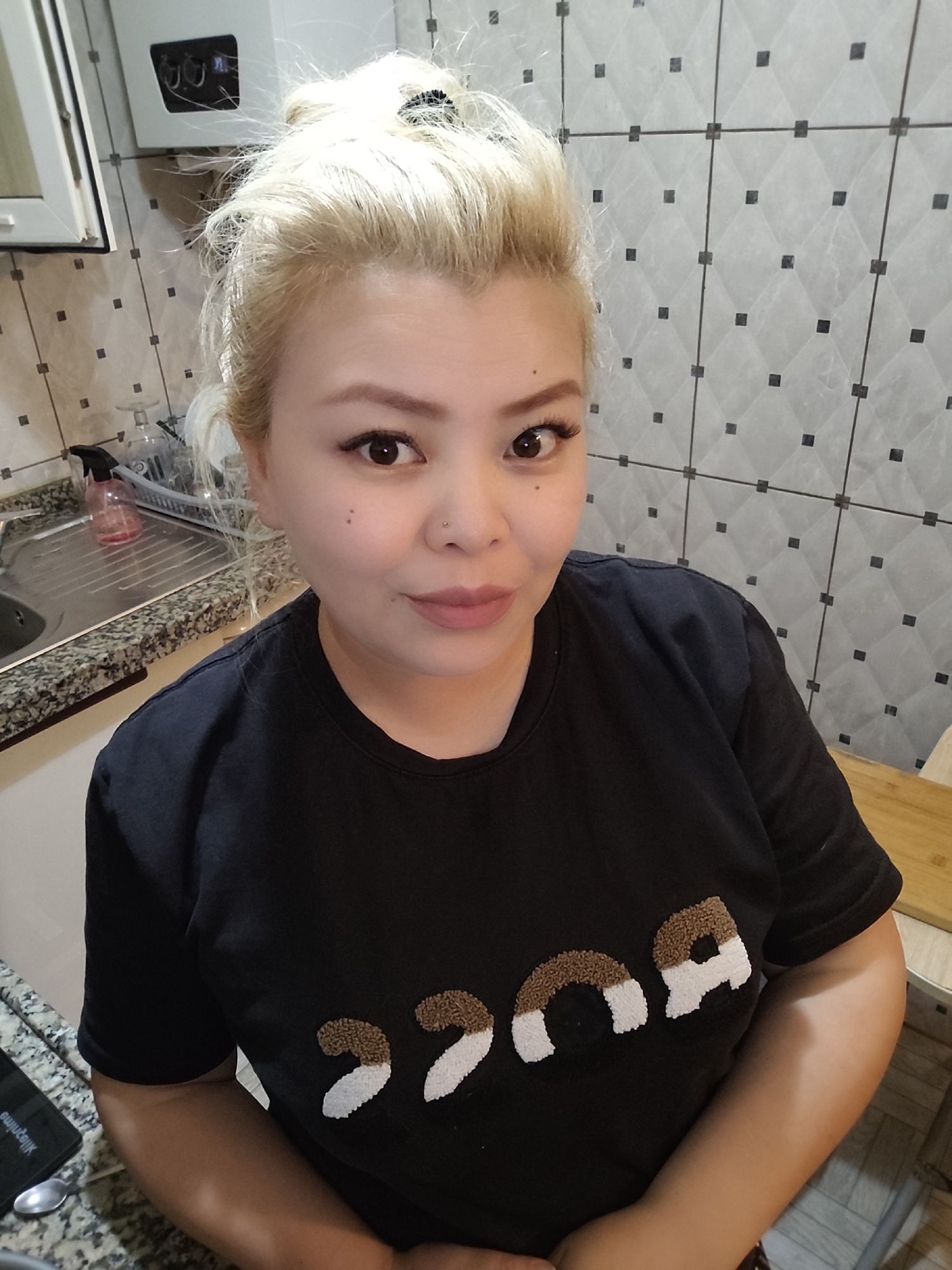 A woman with blonde hair pulled into a high bun stands in a kitchen. She wears a black t-shirt with what appears to be textured lettering across the chest. The kitchen features tiled walls with a geometric pattern, a stainless-steel sink with a granite countertop, and a white appliance on the wall. There are dishes visible in the sink. The location is implied to be in Sarıyer, Turkey.