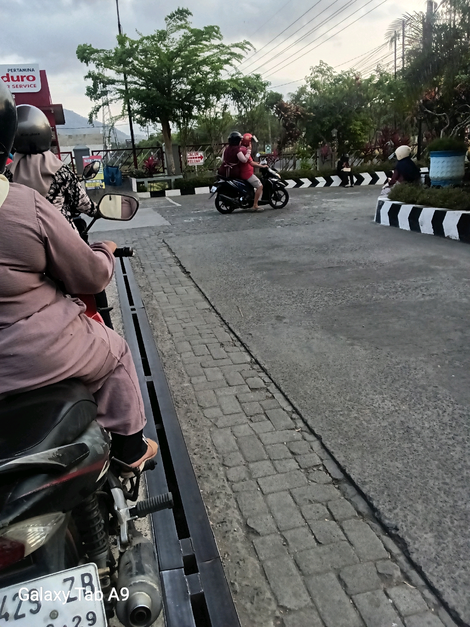 The image captures a street scene in Trenggalek, Indonesia, likely near a gas station given the "Pertamina" signage visible on a red building. In the foreground, a person is seated on a motorcycle, facing away from the camera, with their legs visible wearing a beige hijab and trousers. Another person on a separate motorcycle, wearing a red shirt and helmet, is riding past in the mid-ground, accompanied by a passenger. In the background, lush green trees and some foliage suggest an outdoor environment. The road surface is paved, with a distinct line of black and white chevrons marking a boundary. Power lines are visible crisscrossing the sky, indicating an urban or semi-urban setting. The sky is overcast, suggesting a cloudy day.