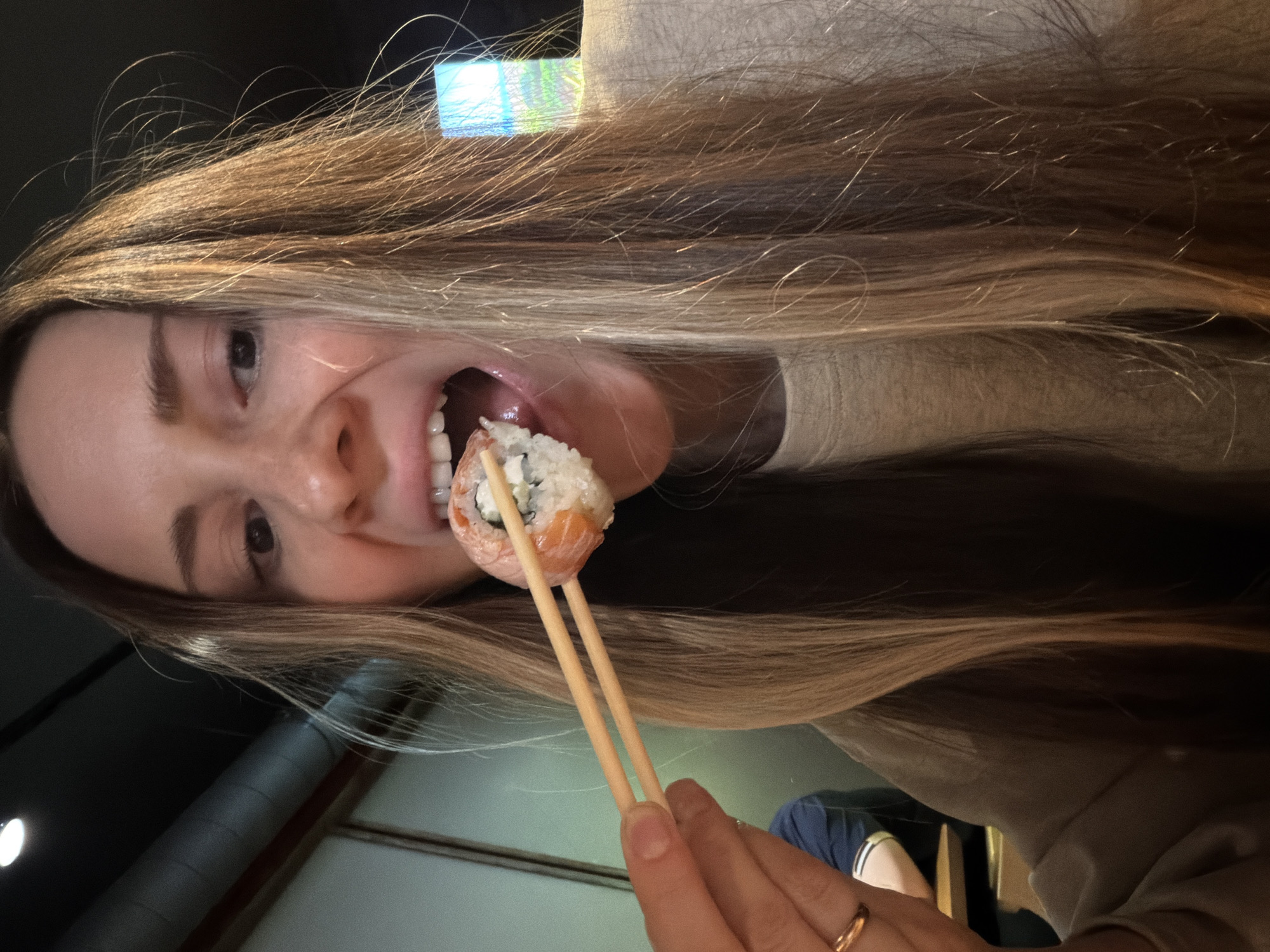 The image captures a close-up, eye-level perspective of a female individual in a restaurant located in Khimki, Russia. The individual, with long, light brown hair, is shown in the active process of consuming a sushi roll. Her mouth is open wide, with a salmon-wrapped sushi roll, containing white rice and a light-colored filling, partially inserted. She holds the sushi roll with light-colored chopsticks in her right hand, on the ring finger of which a gold-colored ring is present. She is wearing a light-colored, possibly beige, long-sleeved garment. In the blurred background to the immediate left, a portion of another person wearing a dark blue shirt is visible. The environment features a dark ceiling with a visible grey pipe or duct and a circular white light fixture. A bright, rectangular light source, exhibiting blue and green tones, is present in the upper right background.