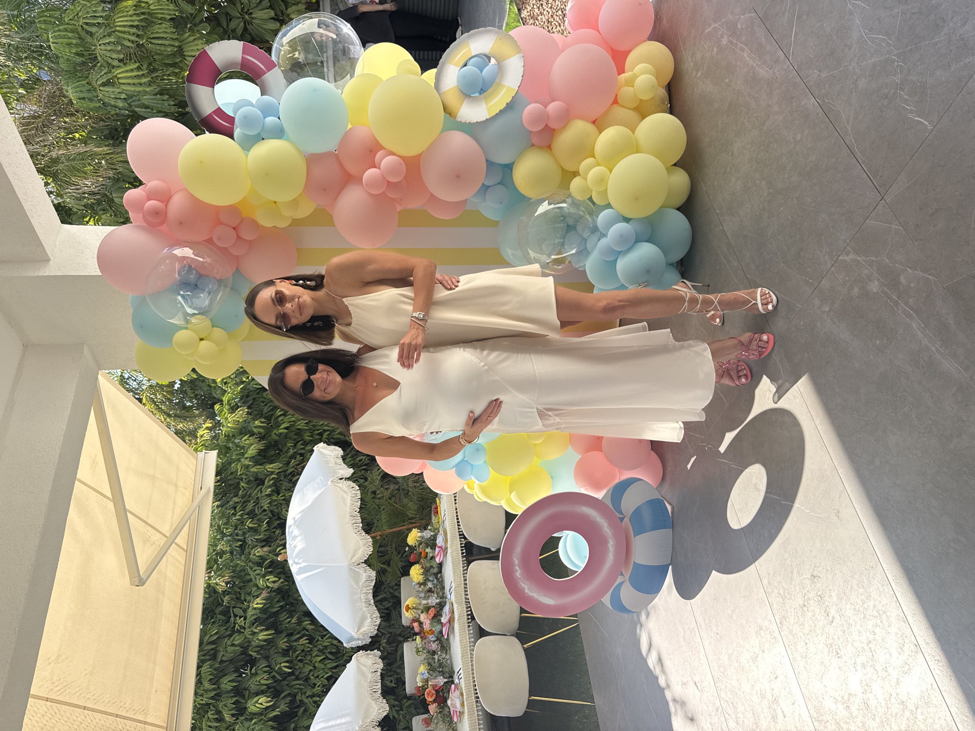 Two women are posing for a photo in an outdoor setting, likely a backyard or patio, with a large balloon arch and a decorated table in the background. The woman on the left is pregnant and wearing a long, white, V-neck dress with lace detailing on the side. She has dark hair and is wearing sunglasses and pink strappy heels. The woman on the right is wearing a short, cream-colored halter dress and white strappy heels. She also has dark hair and is wearing sunglasses.
The balloon arch is a prominent feature, made up of various pastel-colored balloons in shades of pink, yellow, and blue, with some clear balloons mixed in. A decorative life preserver and inflatable pool rings are placed around the balloon arch and in front of the women.
Behind the women, a table is set for a meal with white tablecloths, floral arrangements, and place settings. Two white umbrellas with fringe are also visible. The ground is paved with large, light gray tiles. The background shows lush green foliage and a portion of a building with an awning. The lighting suggests it is daytime and sunny, with distinct shadows cast on the ground.
The scene appears to be a celebration, possibly a baby shower, given the pregnant woman and festive decorations. The location is identified as Agios Tychona Community, Cyprus.