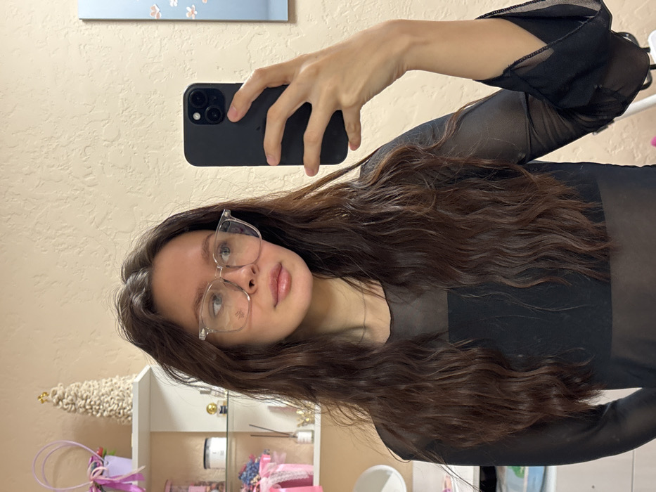 A young woman with long brown wavy hair and clear-framed glasses is holding a black iPhone to take a selfie. She is wearing a black sheer top with long sleeves. The background includes a beige wall with a picture frame and a white cabinet filled with decorative items, including a small Christmas tree. The lighting is soft and natural, creating a casual and intimate atmosphere.
