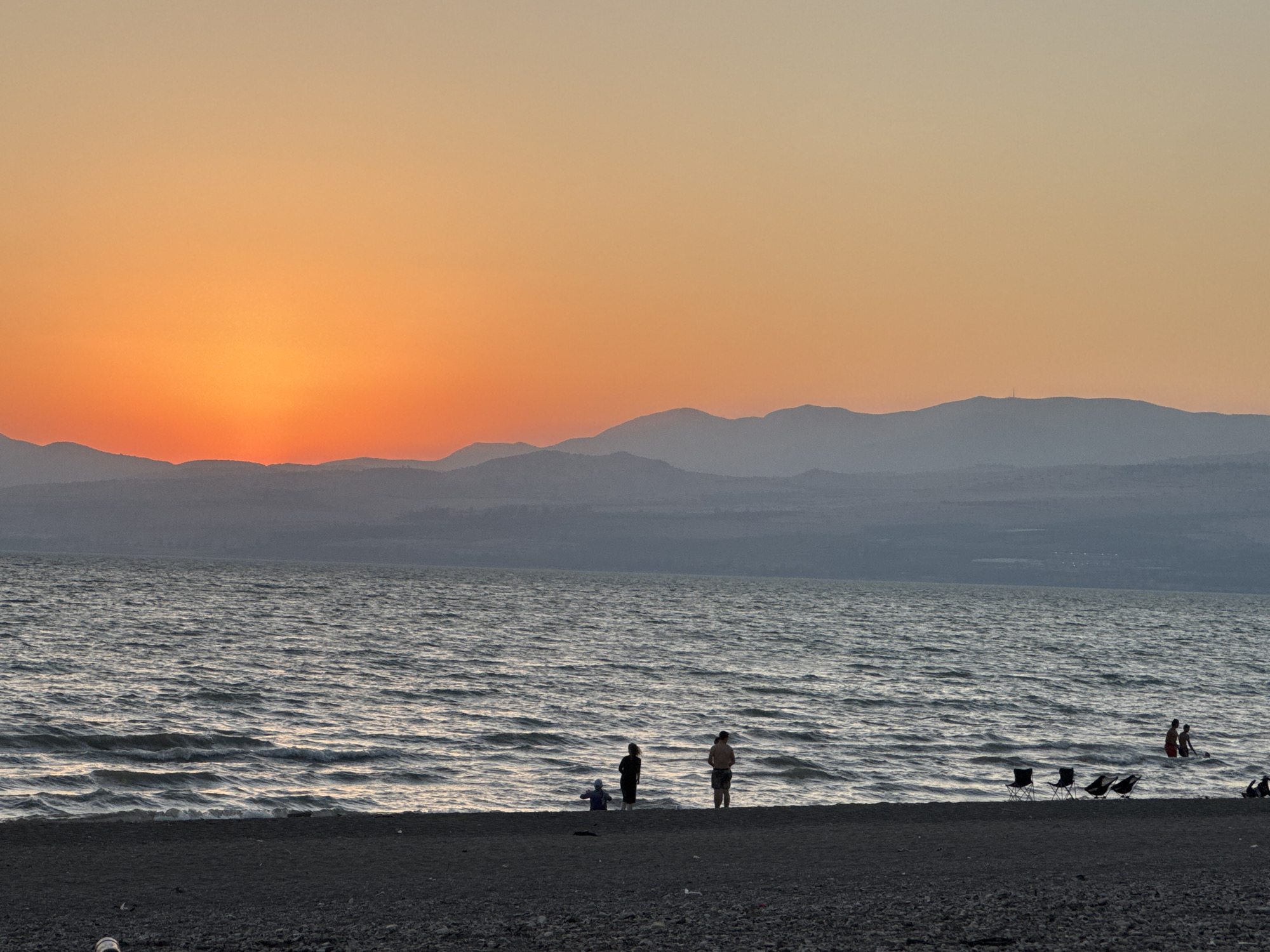The image captures a serene sunset scene at the Sea of Galilee, within the Golan Regional Council, Israel. The sky is ablaze with vibrant orange and yellow hues, transitioning to a soft peach tone as it ascends. Silhouetted against this dramatic sky are distant, hazy mountains. The sea stretches out, its surface rippled by a gentle breeze, reflecting the warm tones of the sunset. On the dark, pebbly shore, several people are visible. In the foreground, a few folding chairs are set up, suggesting a leisurely gathering. Further down the beach, groups of people are scattered. Some are standing near the water's edge, while others are partially submerged in the shallow waves, perhaps cooling off or enjoying the twilight. A lone figure stands apart, looking out at the water. The overall atmosphere is one of tranquility and natural beauty, with the setting sun casting a warm glow over the landscape and its visitors.