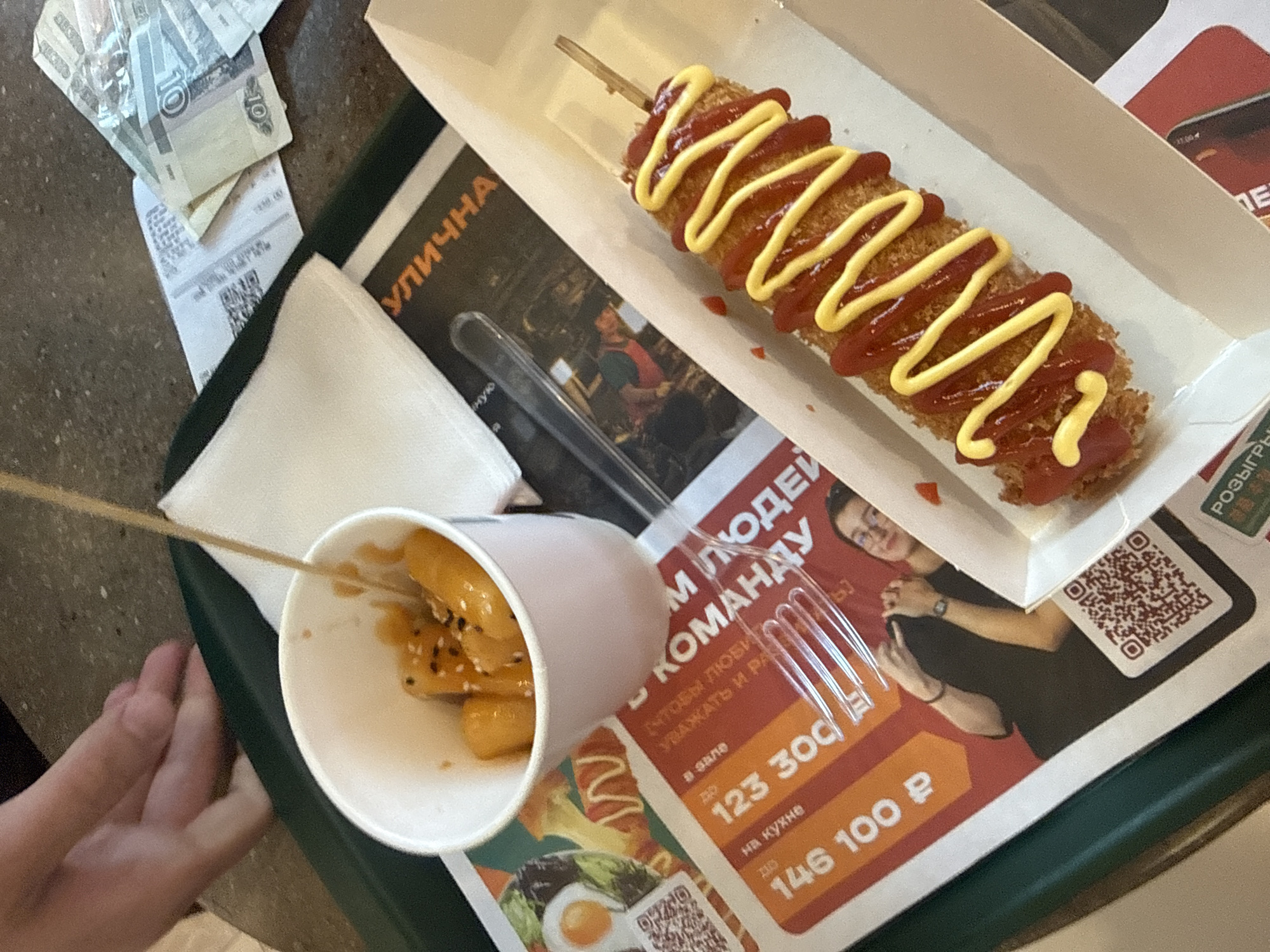 An overhead shot depicts a meal served on a dark green plastic tray, placed on a speckled, light brown tabletop in what is identified as Saint Petersburg, Russia. The scene features various food items, utensils, and printed materials.
In the right foreground, a deep-fried corn dog or Korean hot dog rests in a white cardboard boat. It is generously topped with zigzag patterns of red ketchup and yellow mustard, and a wooden skewer protrudes from its upper end. To the left, a white paper cup holds several orange-yellow, cylindrical or oval-shaped items, likely rice cakes (tteokbokki), coated in a glossy sauce and sprinkled with black sesame seeds. A wooden skewer is also present within this cup.
A clear plastic fork is positioned on a decorative paper placemat, which covers a significant portion of the tray. This placemat features a red and orange design with white Russian text. Visible text includes phrases like "КОМАНДУ" (team), "УЛИЧНА" (street, likely part of "street food"), and financial figures in Rubles such as "123 300 p" and "146 100 p", possibly related to salaries or prizes. The placemat also displays images, including an illustration of a meal with an egg and seaweed, a photograph of a young person in glasses making a hand gesture, and two QR codes.
To the upper right of the tray, on the tabletop, several Russian Ruble banknotes are scattered alongside white paper receipts, one of which also contains a QR code. A 10 Ruble banknote is clearly visible. In the upper left corner of the frame, a fair-skinned human hand is partially visible, resting on the edge of the tray or table, suggesting the presence of an individual interacting with the meal.