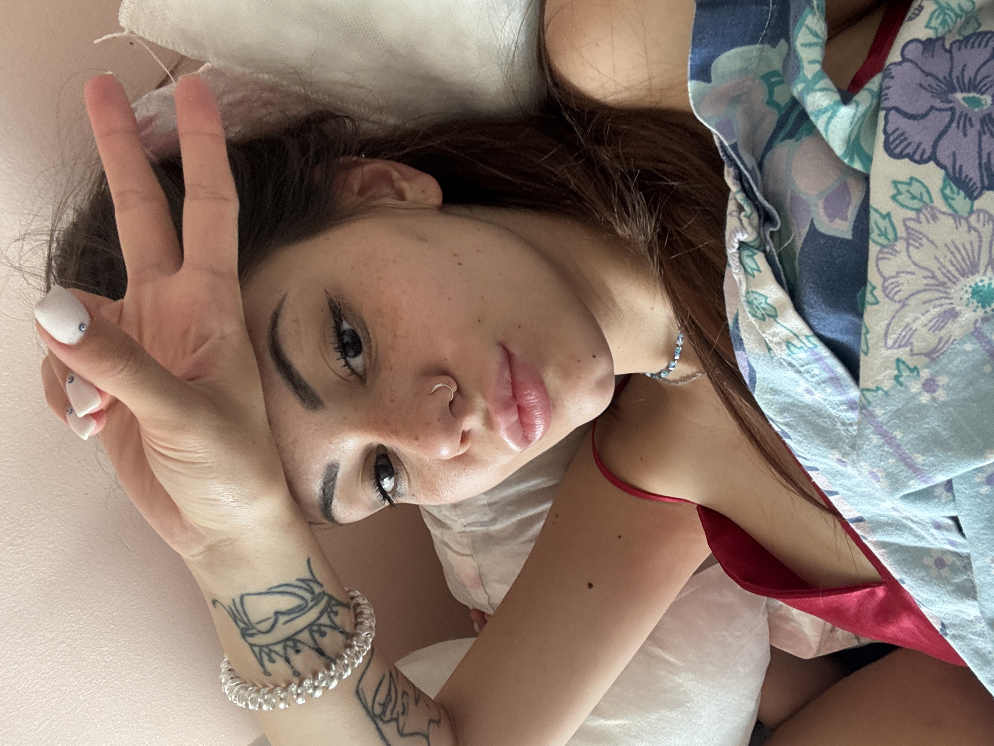 A young woman is captured in a close-up, high-angle shot, lying down indoors, likely in a bed or on a soft surface. She gazes directly at the camera with a soft, slightly alluring expression. Her long, dark brown hair frames her face, which is dotted with freckles. She has pronounced eyebrows and eyeliner, and a silver hoop nose ring on her left nostril. Her lips are full and appear glossy.
Her left hand is raised, forming a peace sign (V-sign) with her index and middle fingers, partially obscuring her forehead. Her fingernails are painted white, with small rhinestone details visible on her thumb. A silver coiled bracelet adorns her left wrist, and a tattoo, resembling an ornate crown or intricate design, is visible on her left forearm.
She is dressed in a red or deep pink camisole or slip, partially covered by a loosely draped outer garment. This garment features a vibrant floral pattern in shades of blue, purple, and green. The background is simple and out of focus, consisting of light-colored bedding and a light-toned wall. The lighting suggests daytime. The scene is intimate and casual, set in Sofia, Bulgaria. No text is visible in the image.