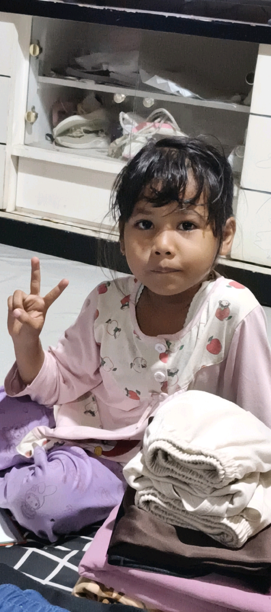 A young Indonesian girl of approximately five years of age is seated on a patterned floor. She is wearing a pink pajama set with a white collar adorned with a red apple print. The girl is facing the camera, her gaze directed forward. Her left hand is raised, forming a "V" sign with her index and middle fingers. In the background, a white cabinet with glass doors contains various items, including what appears to be folded clothing and bags. To the right of the girl, a stack of folded clothes, predominantly in beige and brown hues, is visible. The scene suggests an indoor domestic setting, likely within Muaro Jambi, Indonesia.
