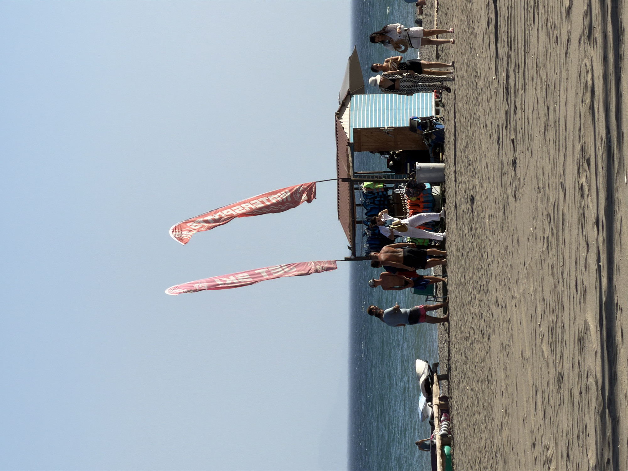 On a sandy beach, several people are gathered near a small stand selling water sports equipment. The stand is open-air with a roof and a blue and white striped wall. Two large red flags with white lettering are prominently displayed, likely advertising the establishment. In the background, the ocean stretches to the horizon under a clear sky. A group of men stand together to the left of the stand, some in swimwear. To the right, three women are interacting near the stand, one in a patterned outfit, another in a black bikini, and a third in a white outfit. Further left, a person is sitting on a low structure by the water, next to some water sports equipment. The overall scene depicts a sunny day at a beach resort.